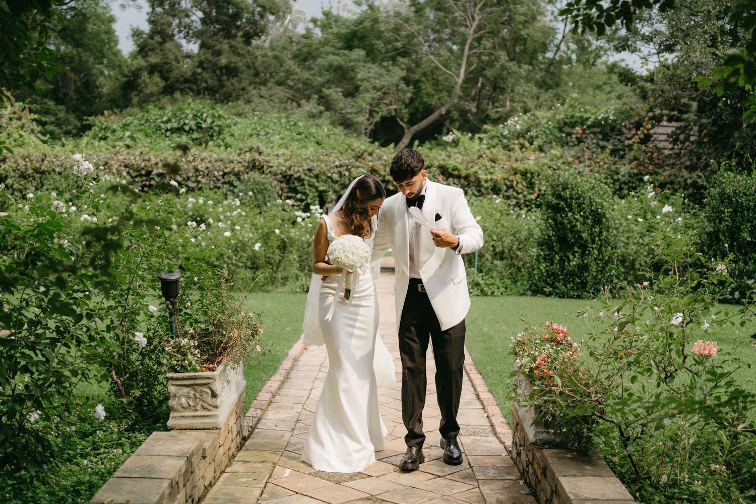 Newlyweds walking together hand in hand on a stone path surrounded by blooming garden flowers.