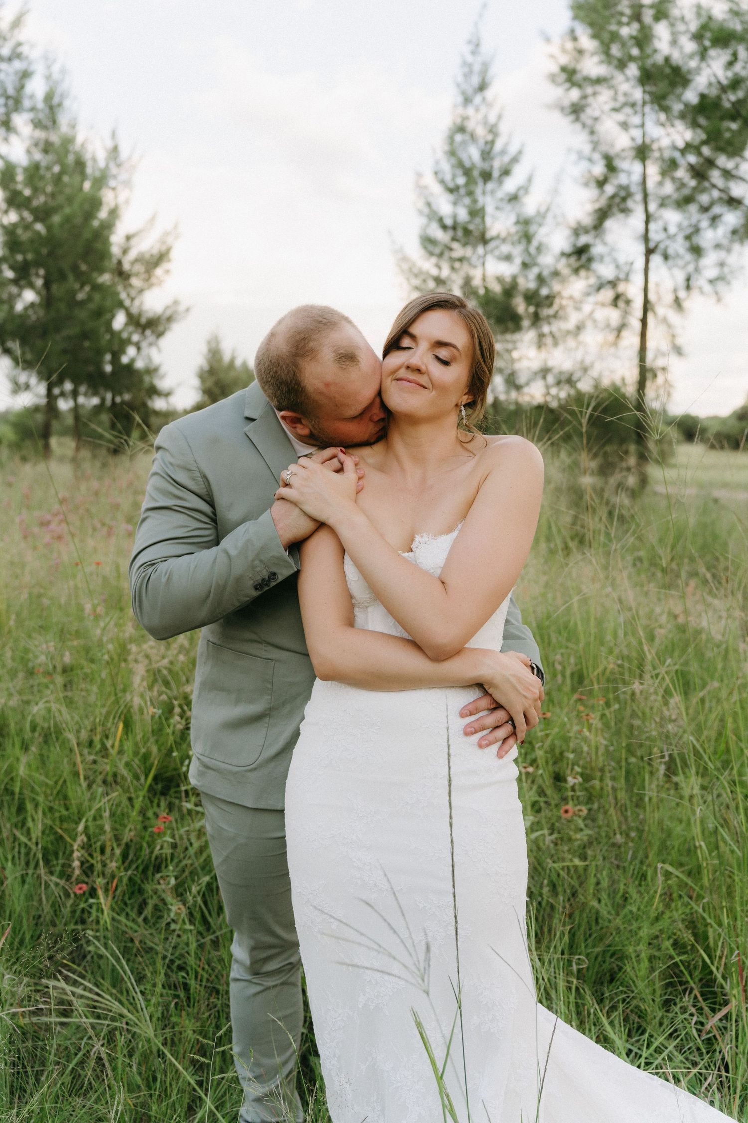 A romantic wedding couple embraces in a sun-lit meadow with pine trees in the background.