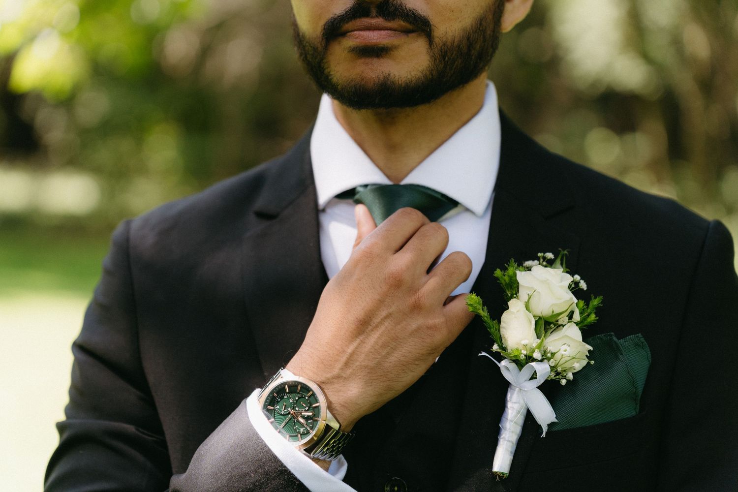 A groom in a black suit adjusts his tie, wearing a white boutonniere and silver watch in an outdoor setting.