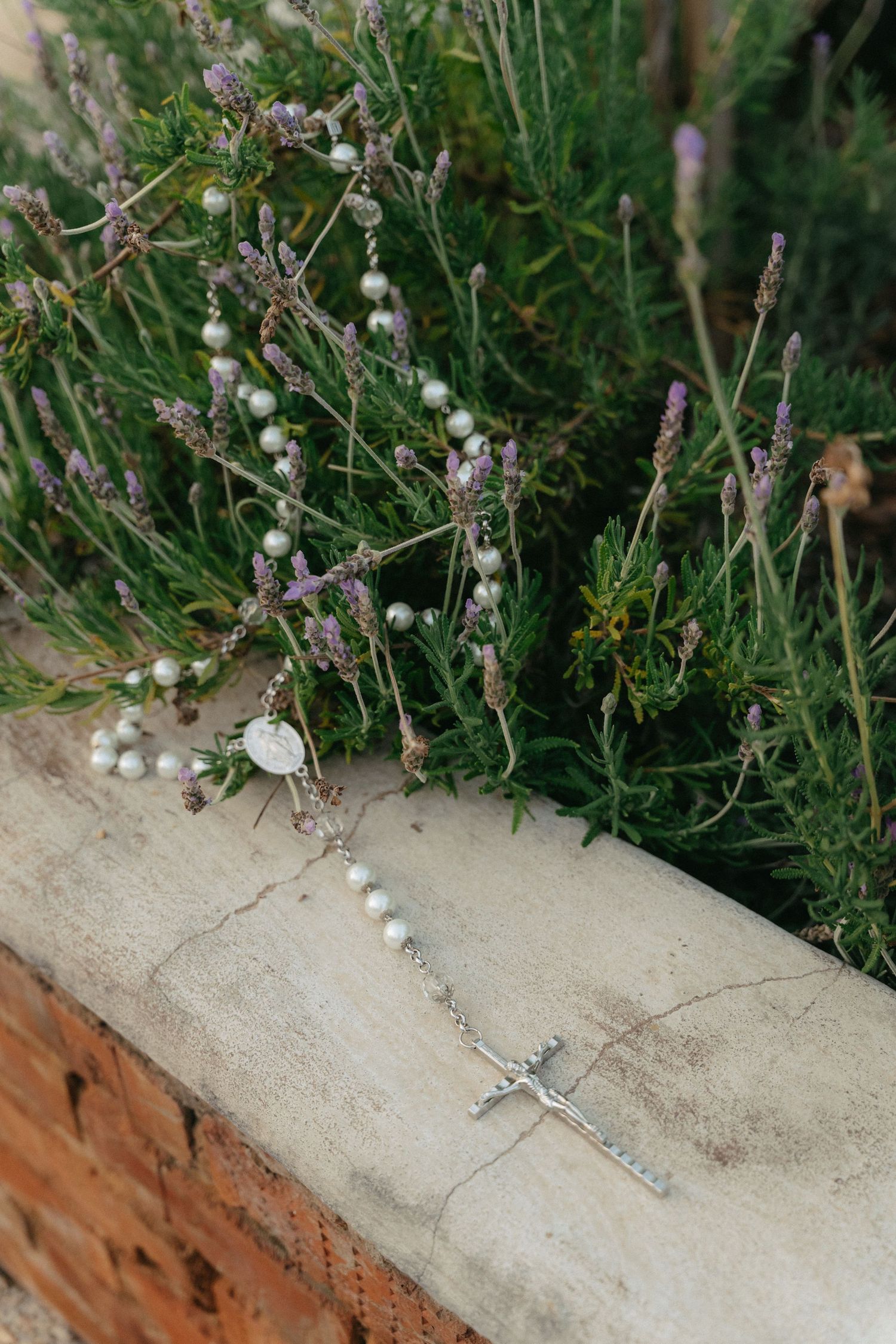 Small silver cross necklace lying among delicate white and purple wildflowers on concrete.