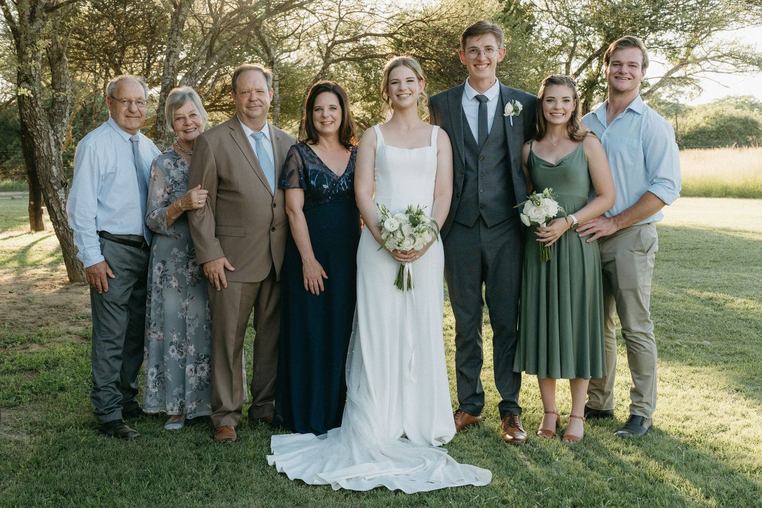 Small wedding party in formal attire poses together outdoors on green lawn in afternoon sunlight.
