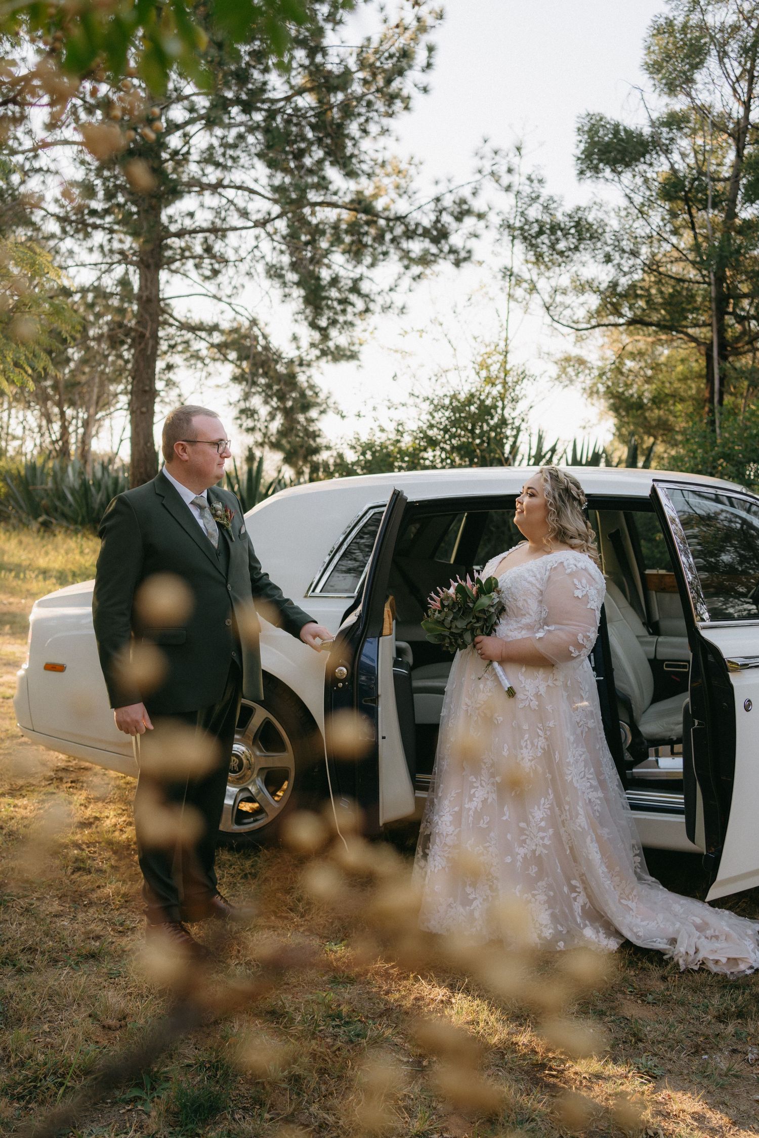 Bride in lace gown and groom stand beside white limousine in sunlit wooded setting.