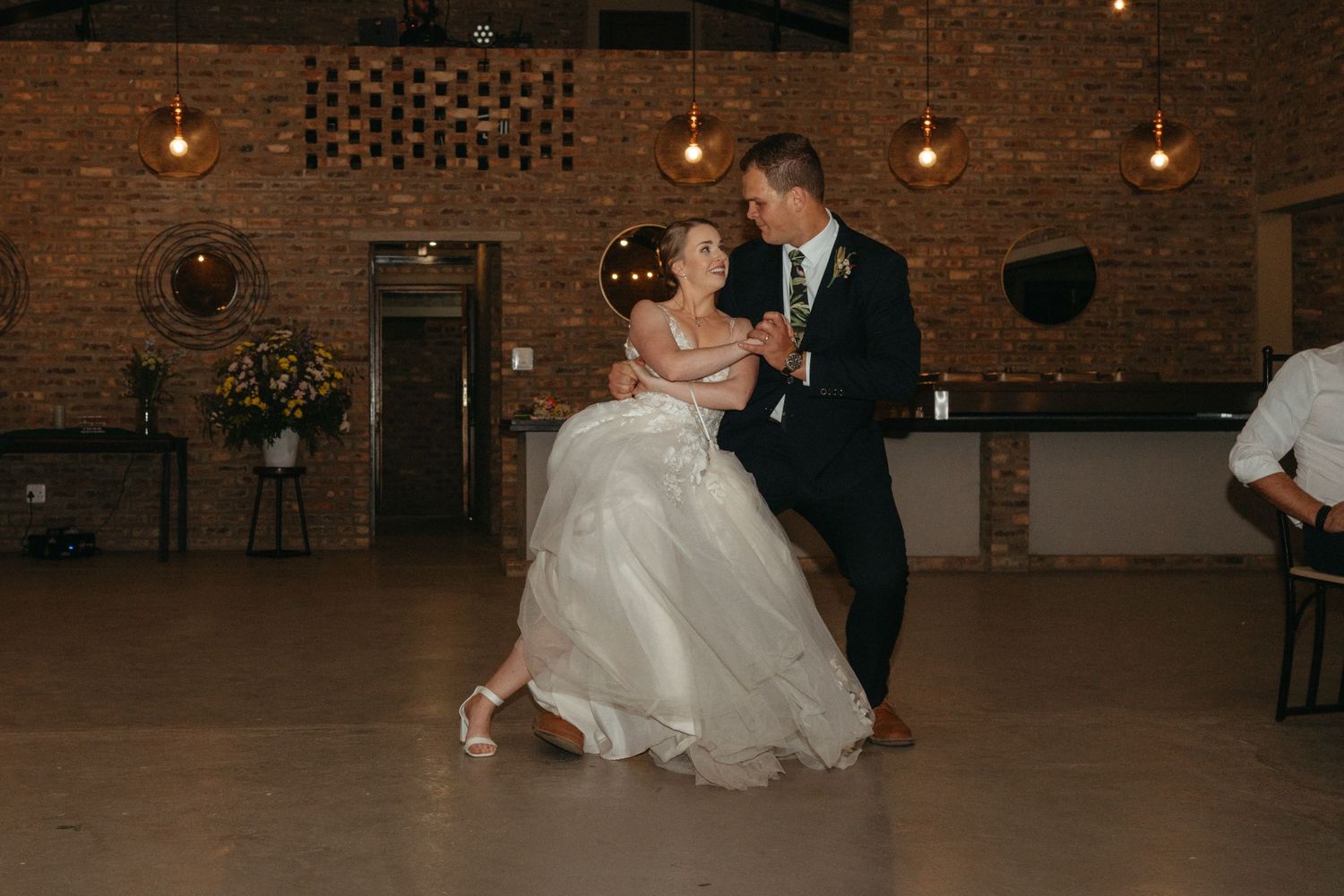 Romantic first dance in a rustic brick venue illuminated by warm overhead lighting.