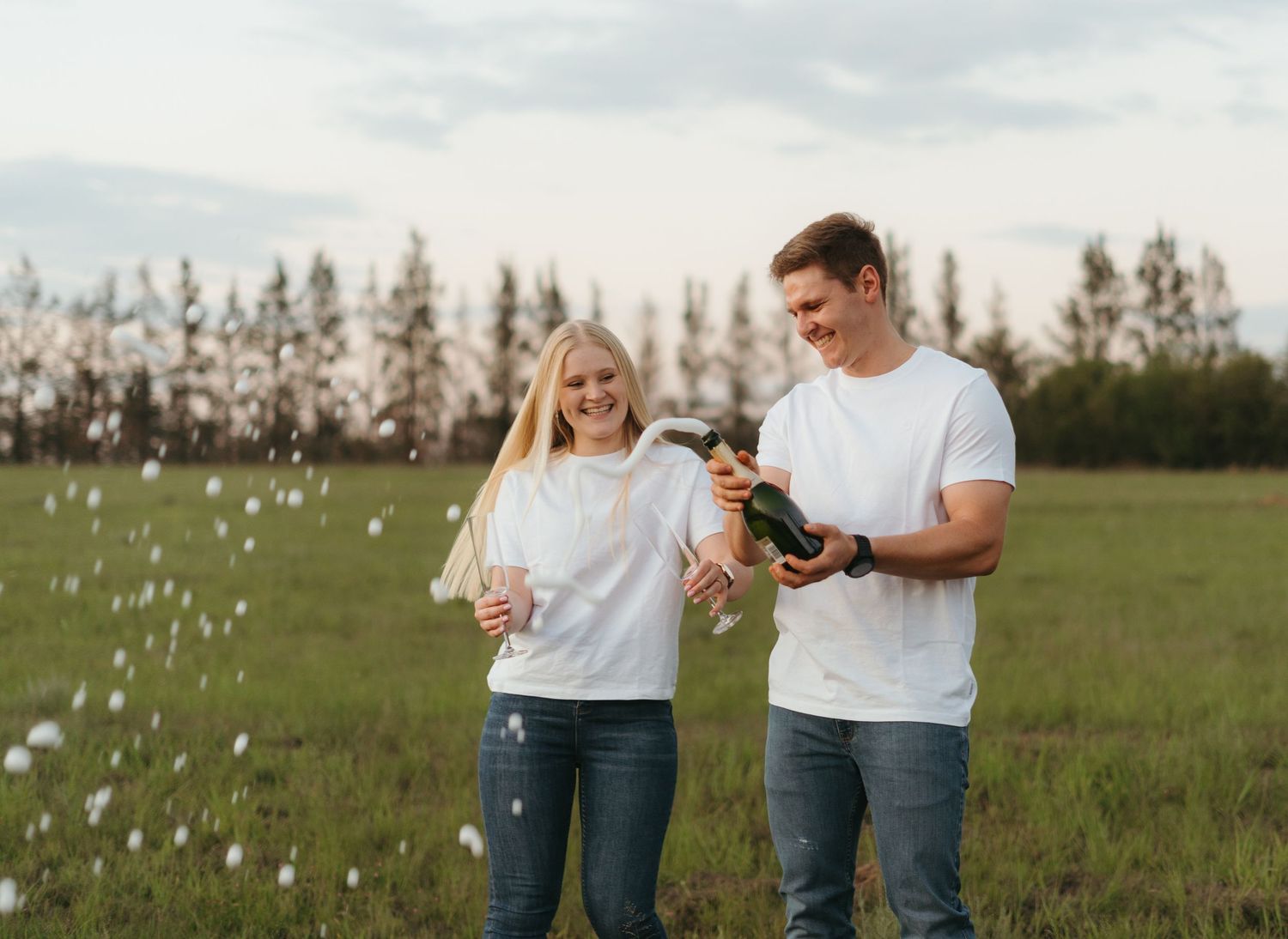 Couple popping champagne in a sunny field with trees in background.