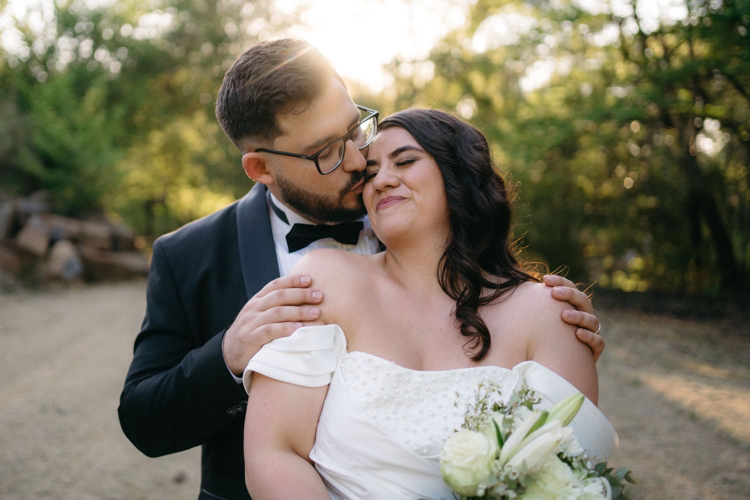 A loving couple embraces outdoors on their wedding day, with the bride in white holding a bouquet of flowers.