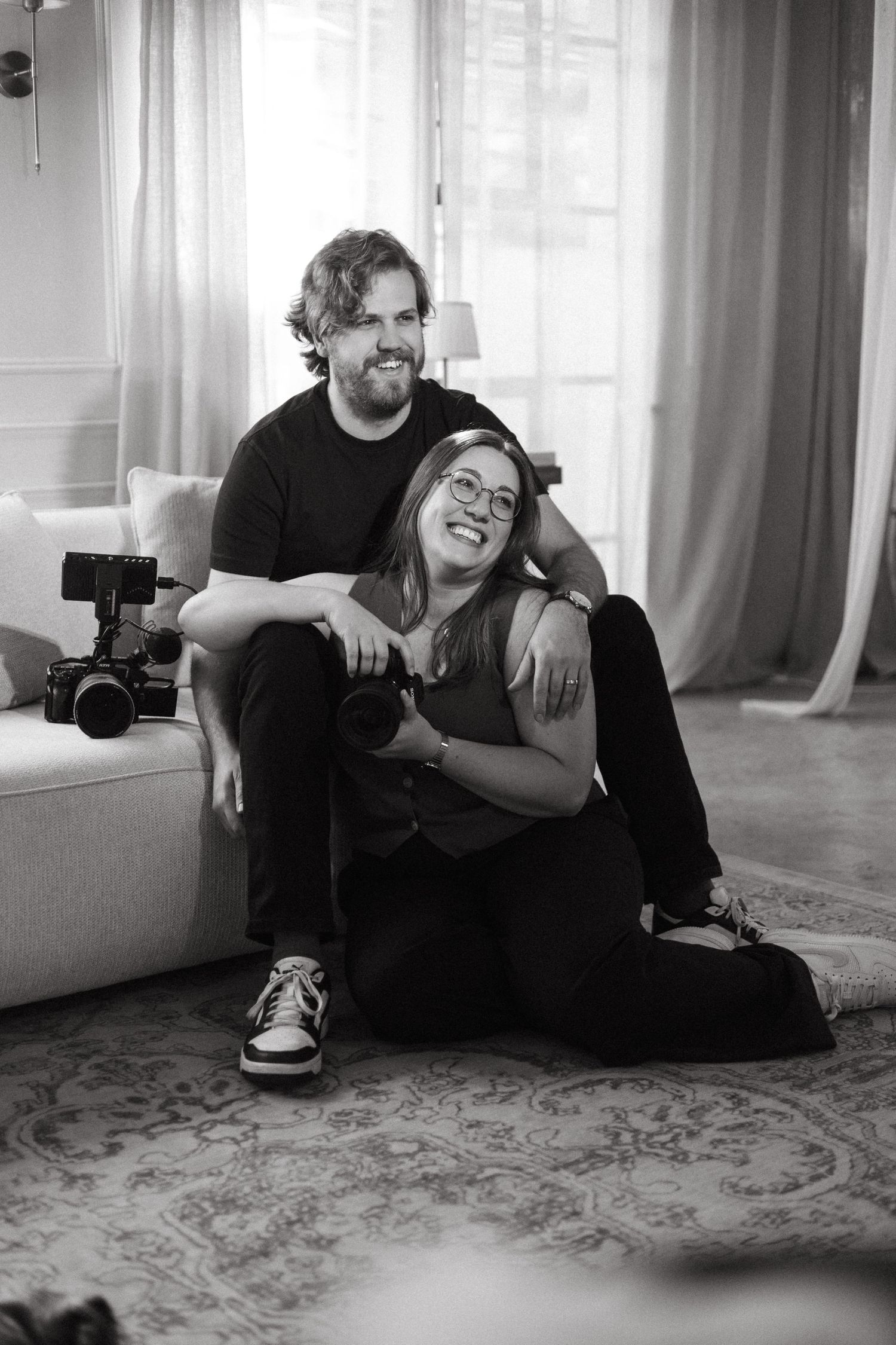 Black and white portrait of a couple sitting on the floor, smiling, holding cameras near a couch in Pretoria, South Africa