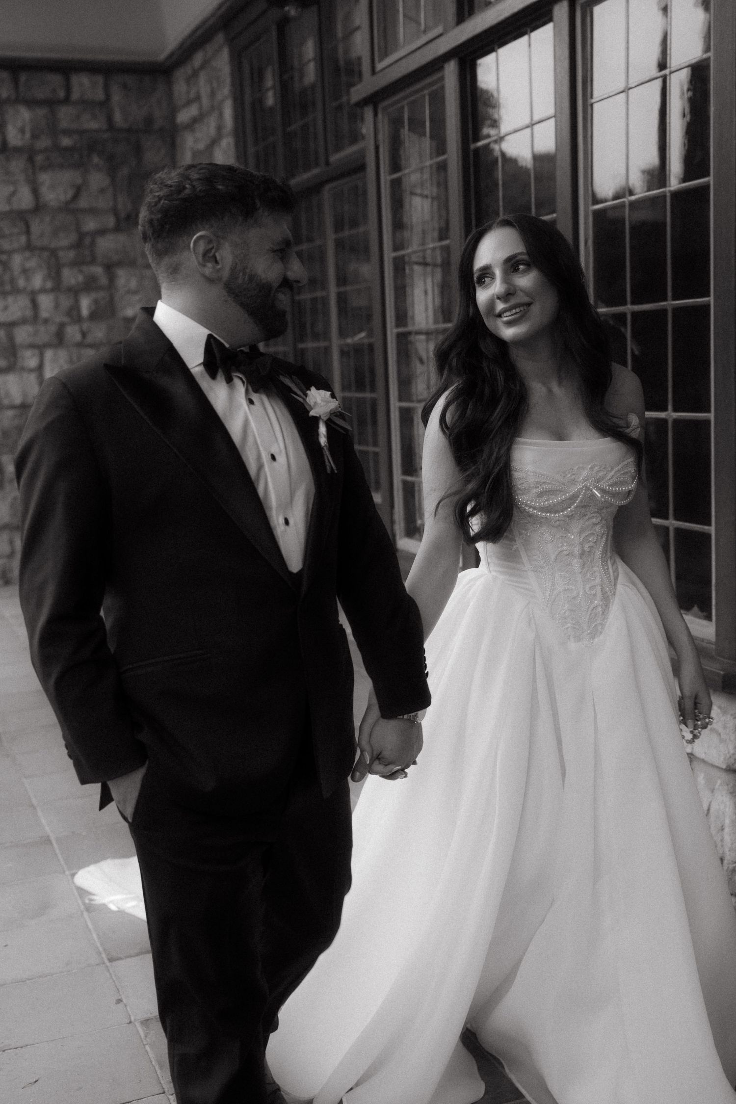 Black and white photo of a bride in a corset gown and groom in a tuxedo holding hands at their wedding.