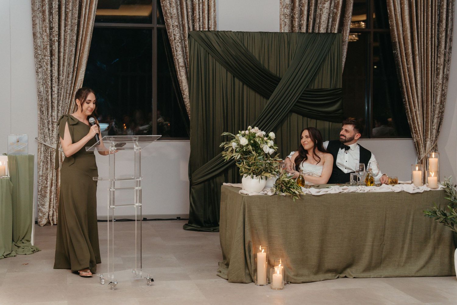 Bridesmaid gives speech at wedding reception while bride and groom sit at decorated sweetheart table with candles.