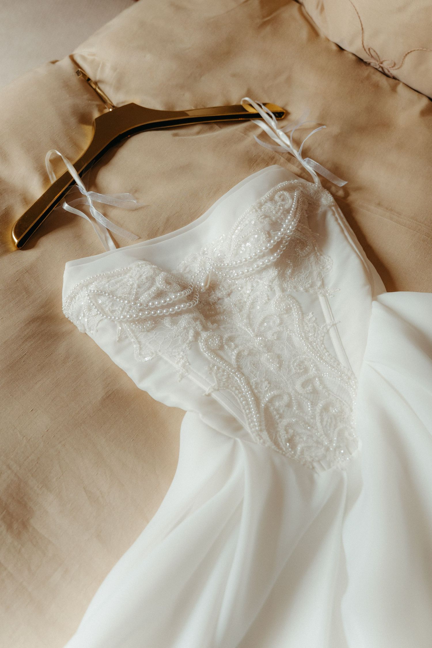 Elegant white lace wedding dress with spaghetti straps on a gold hanger, displayed on beige fabric backdrop.