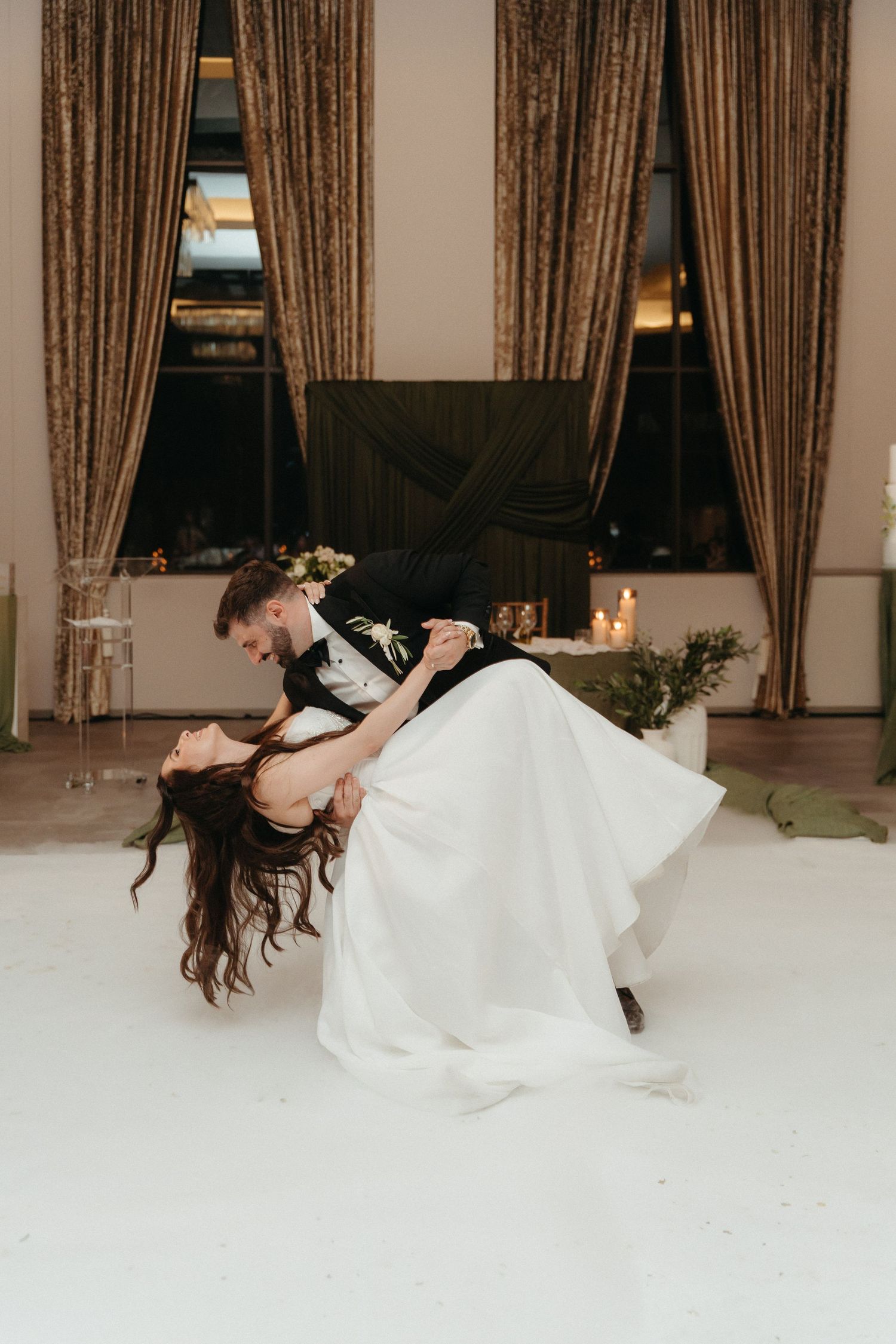 Bride and groom share romantic first dance dip on white dance floor with gold curtains and candlelit backdrop.