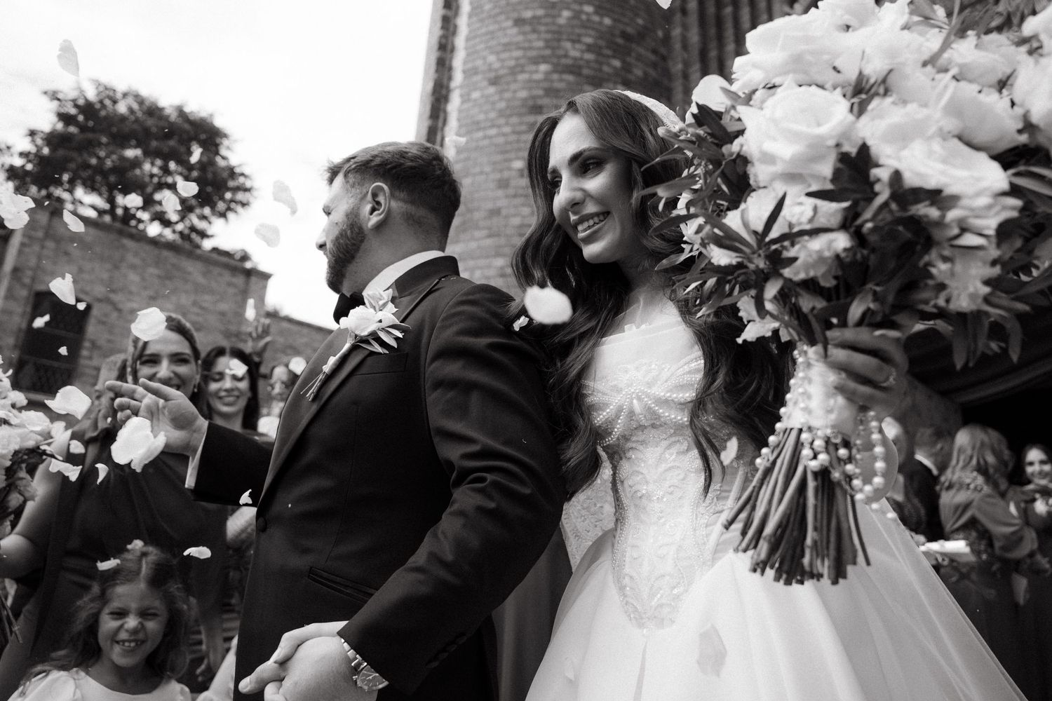 Black and white wedding photo of smiling bride holding white bouquet and groom walking out of Rosebank Catholic Church celebrating.