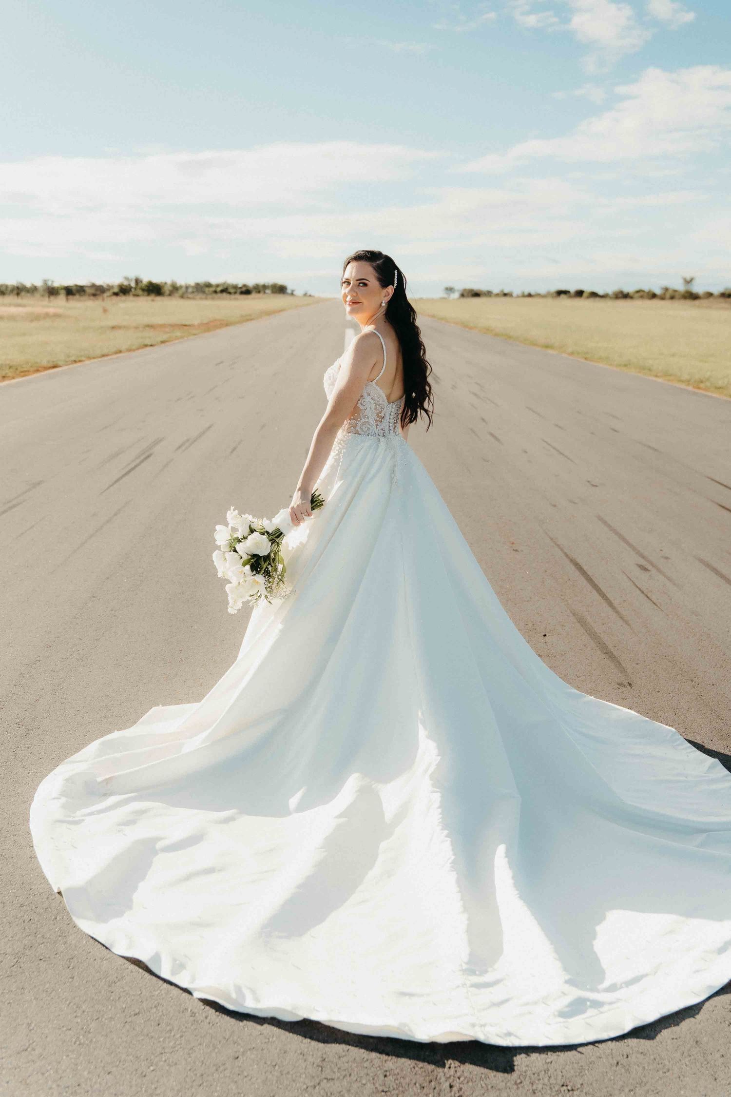 Bride in a flowing white ball gown with long train holds bouquet on open road under blue sky.