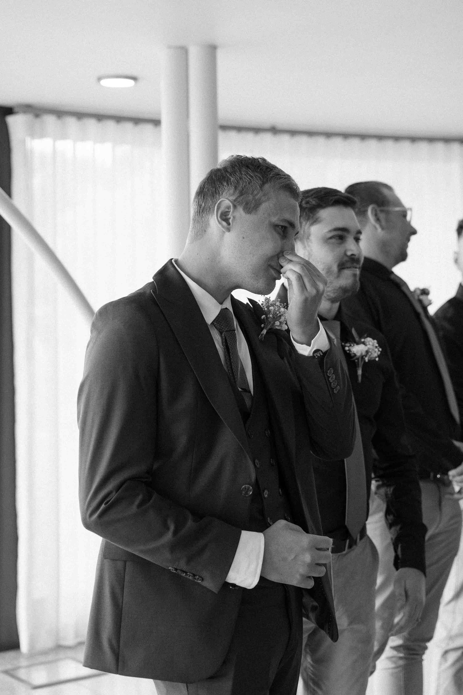 Black and white photo of an emotional groom in a suit wiping tears at his wedding ceremony with groomsmen standing behind.
