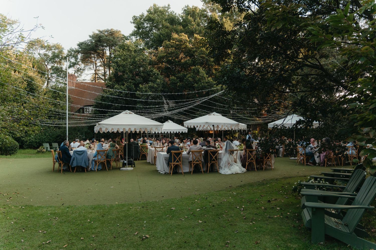 Beechwood Gardens outdoor wedding reception under white canopy tents on lush green lawn surrounded by trees with string lights.
