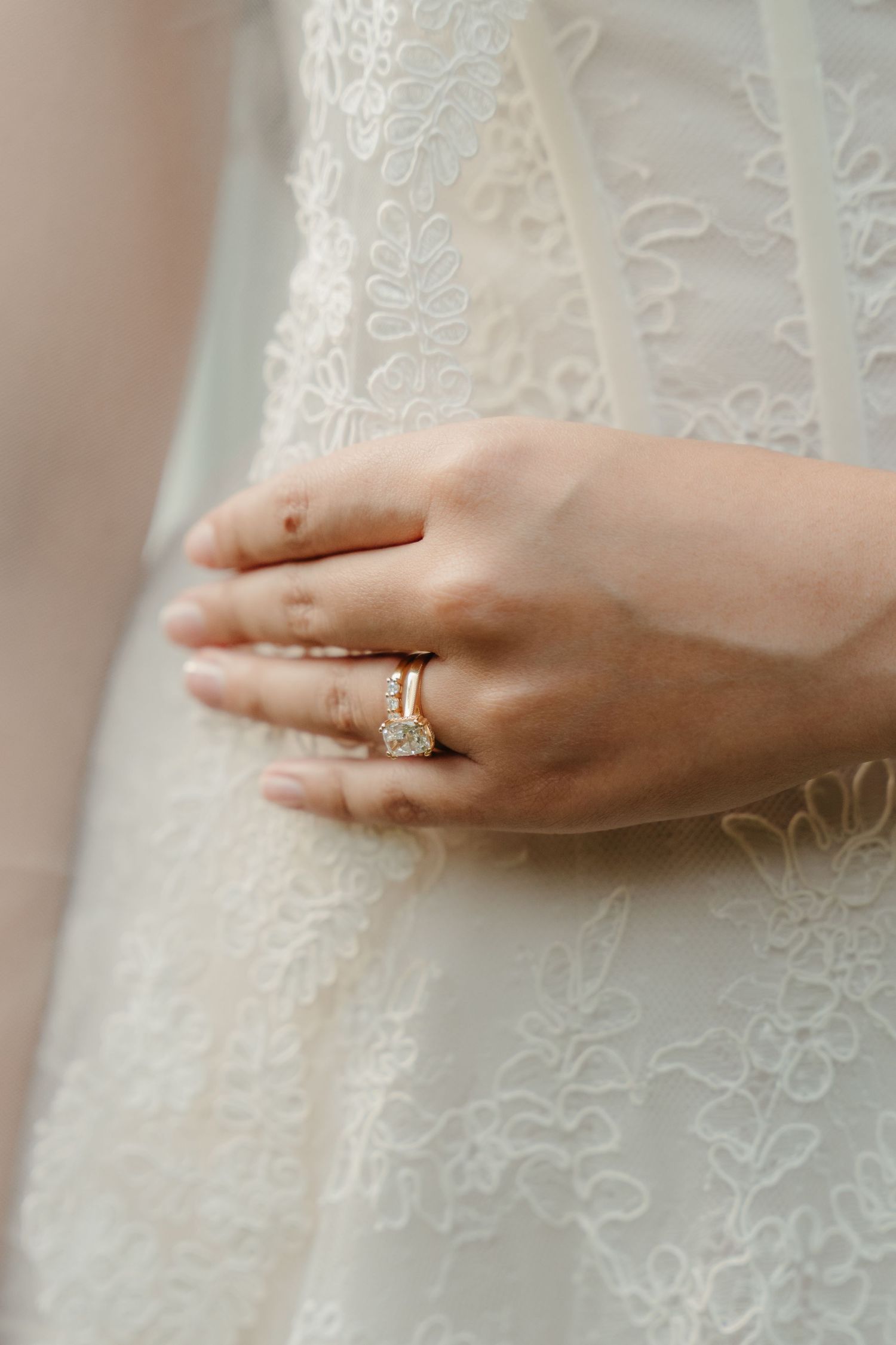 Close-up of a bride's hand wearing a diamond engagement ring and wedding band on a white lace wedding dress.
