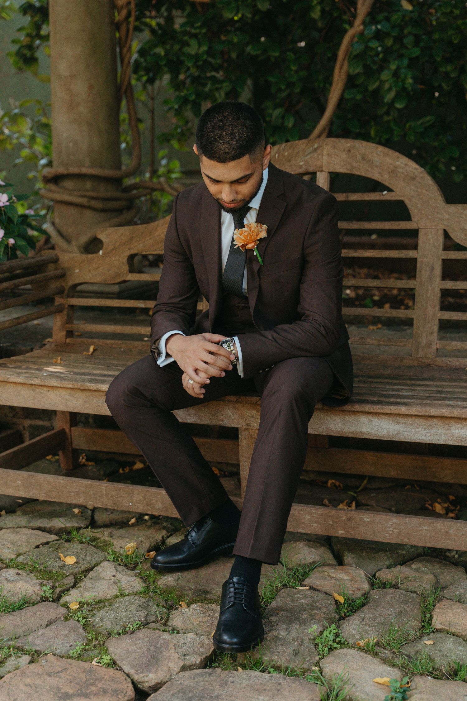 A groom in a brown suit with an orange boutonniere sits thoughtfully on a rustic wooden bench in a garden setting.