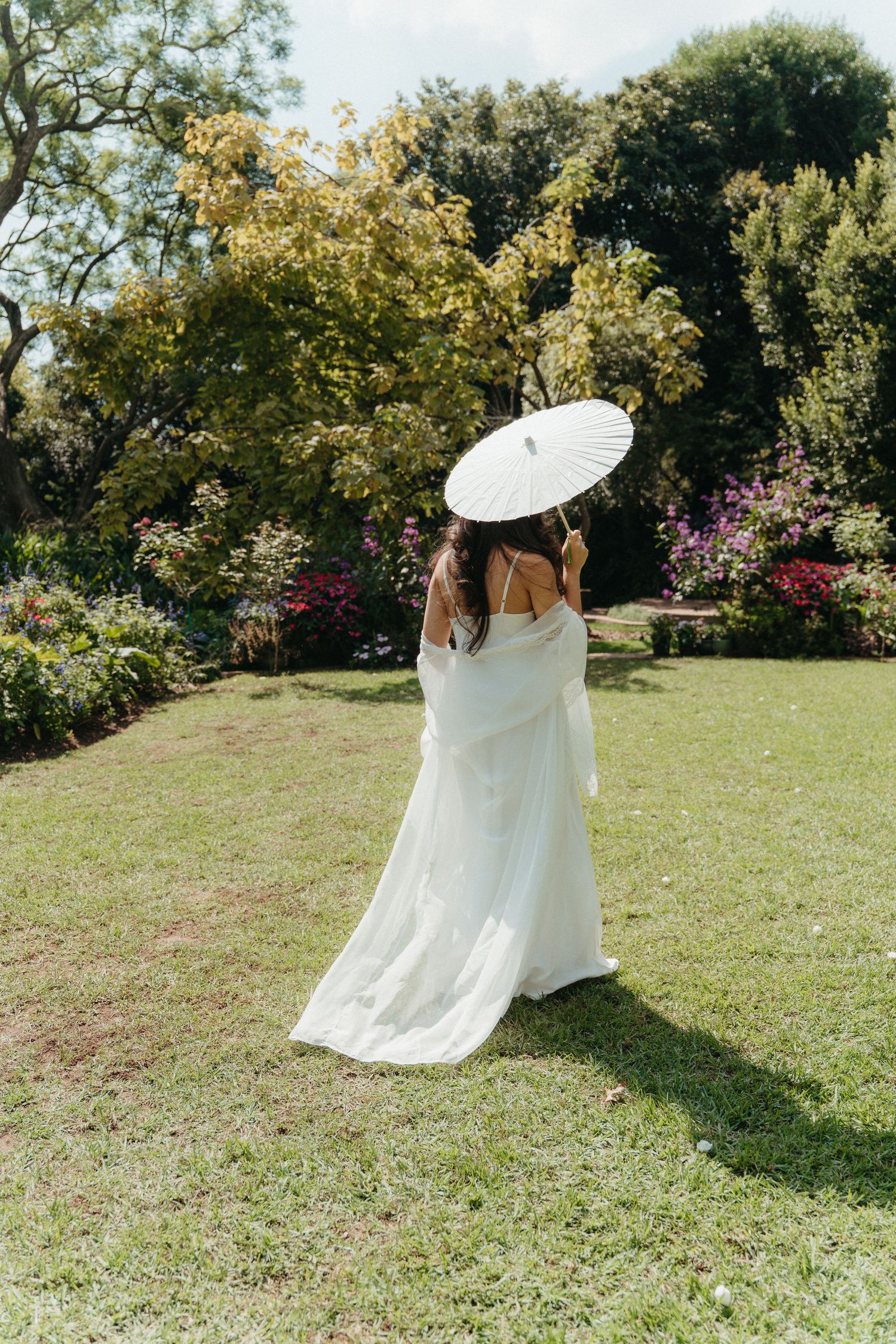 Bride in white dress holding parasol umbrella stands in lush garden with colorful flowers on sunny day.