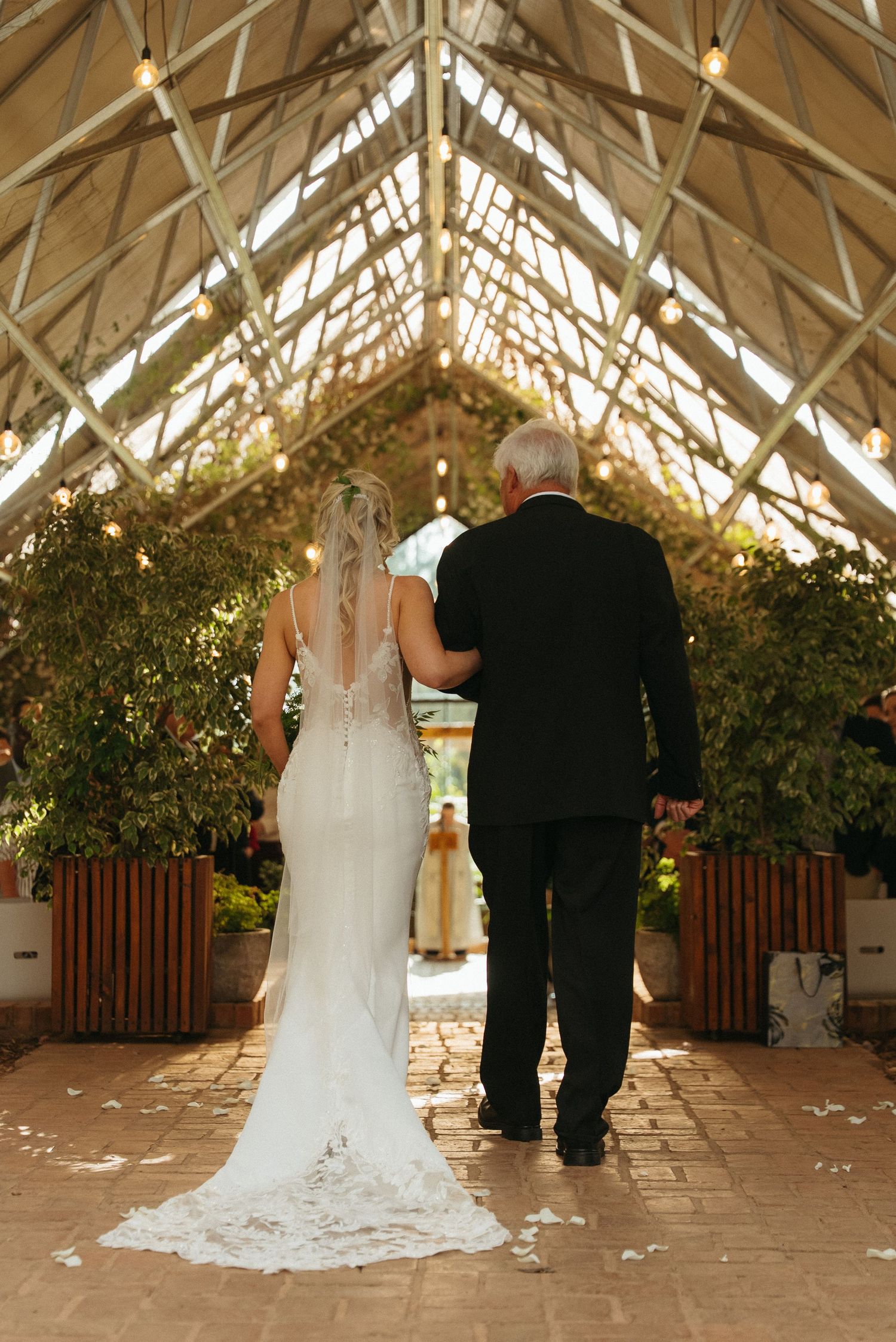 Bride in white gown with veil walks arm-in-arm with older man in black suit down greenhouse aisle.