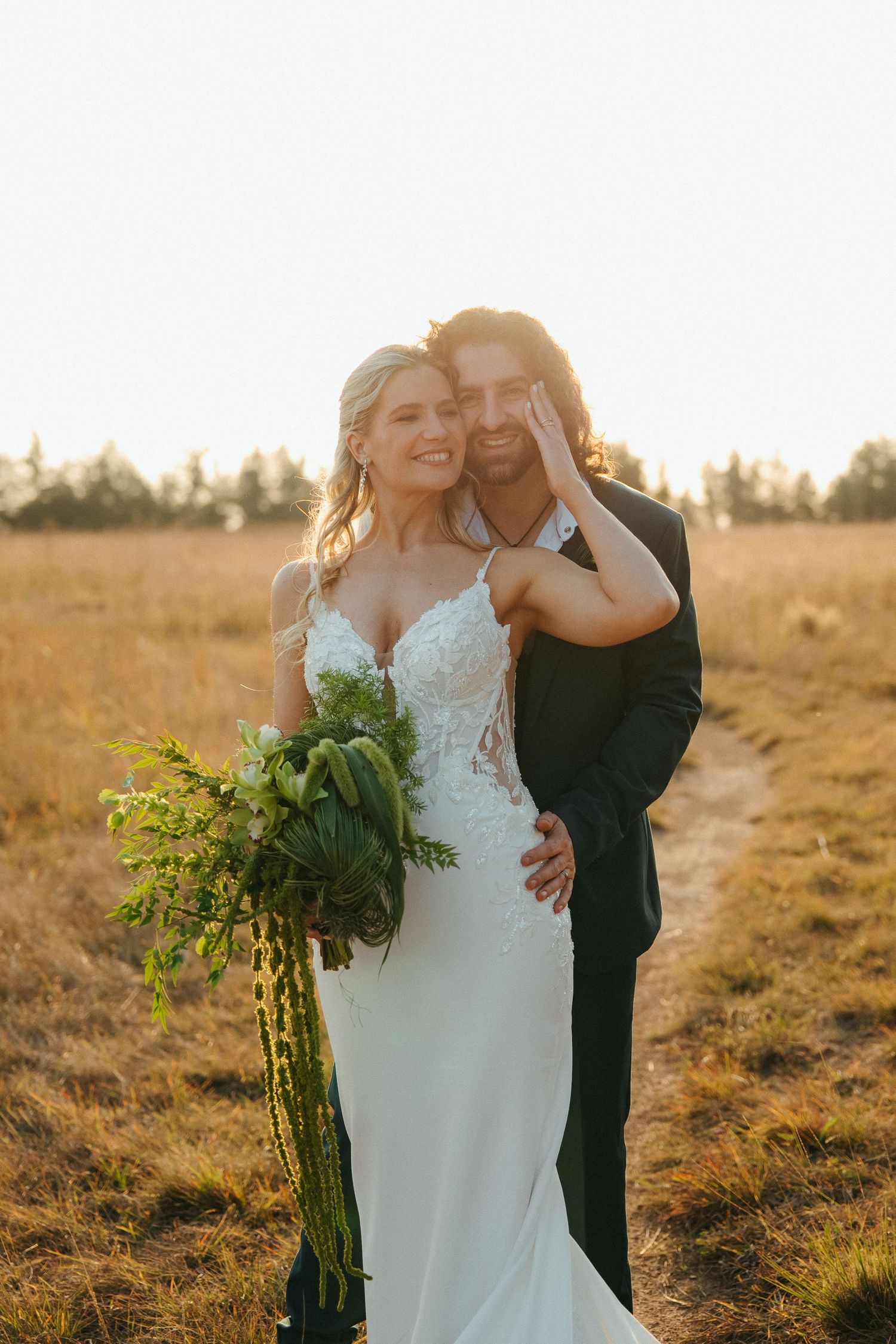 Bride and groom embracing in golden field at sunset, bride holding green bouquet in white lace gown.