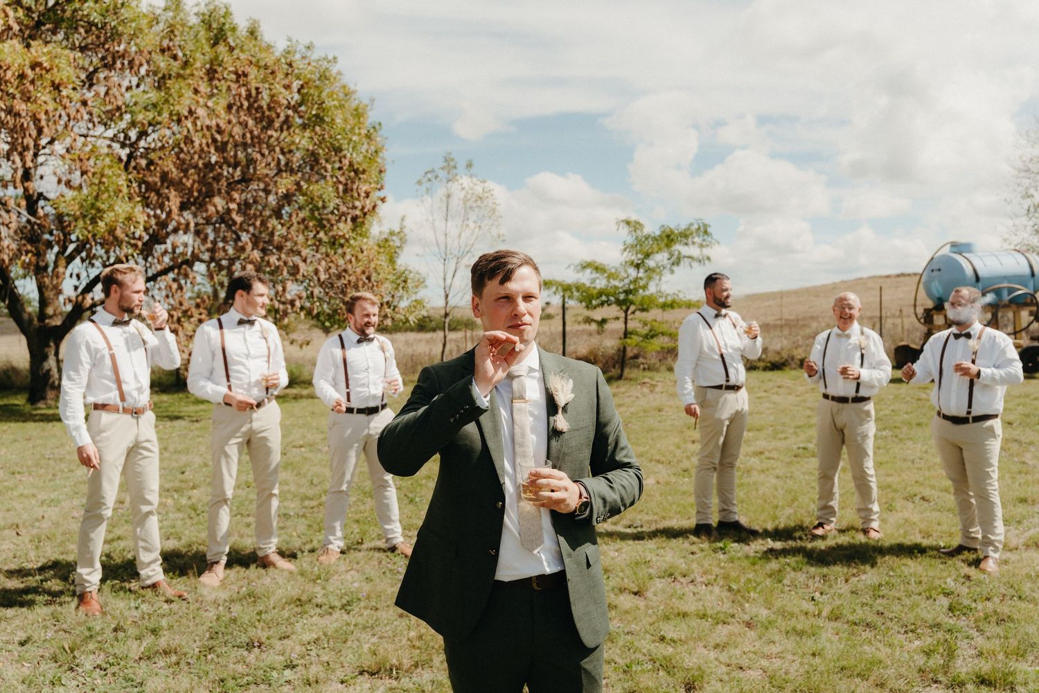 Groom in green suit adjusts tie while groomsmen in white shirts and suspenders stand behind him outdoors.