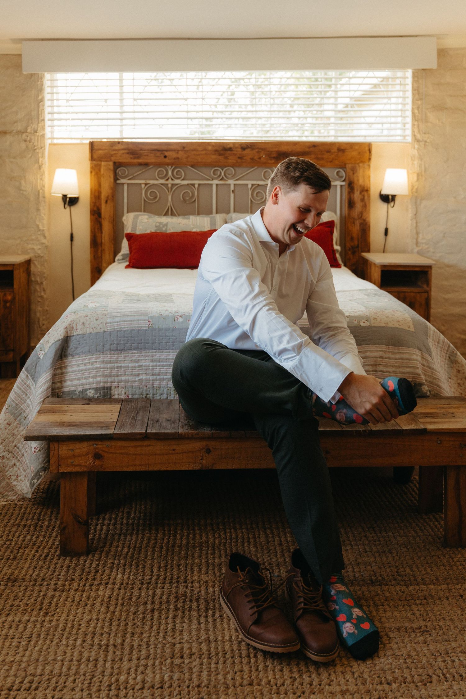 Man sitting on wooden bed bench, smiling while putting on colorful socks, wearing white shirt and dark pants.