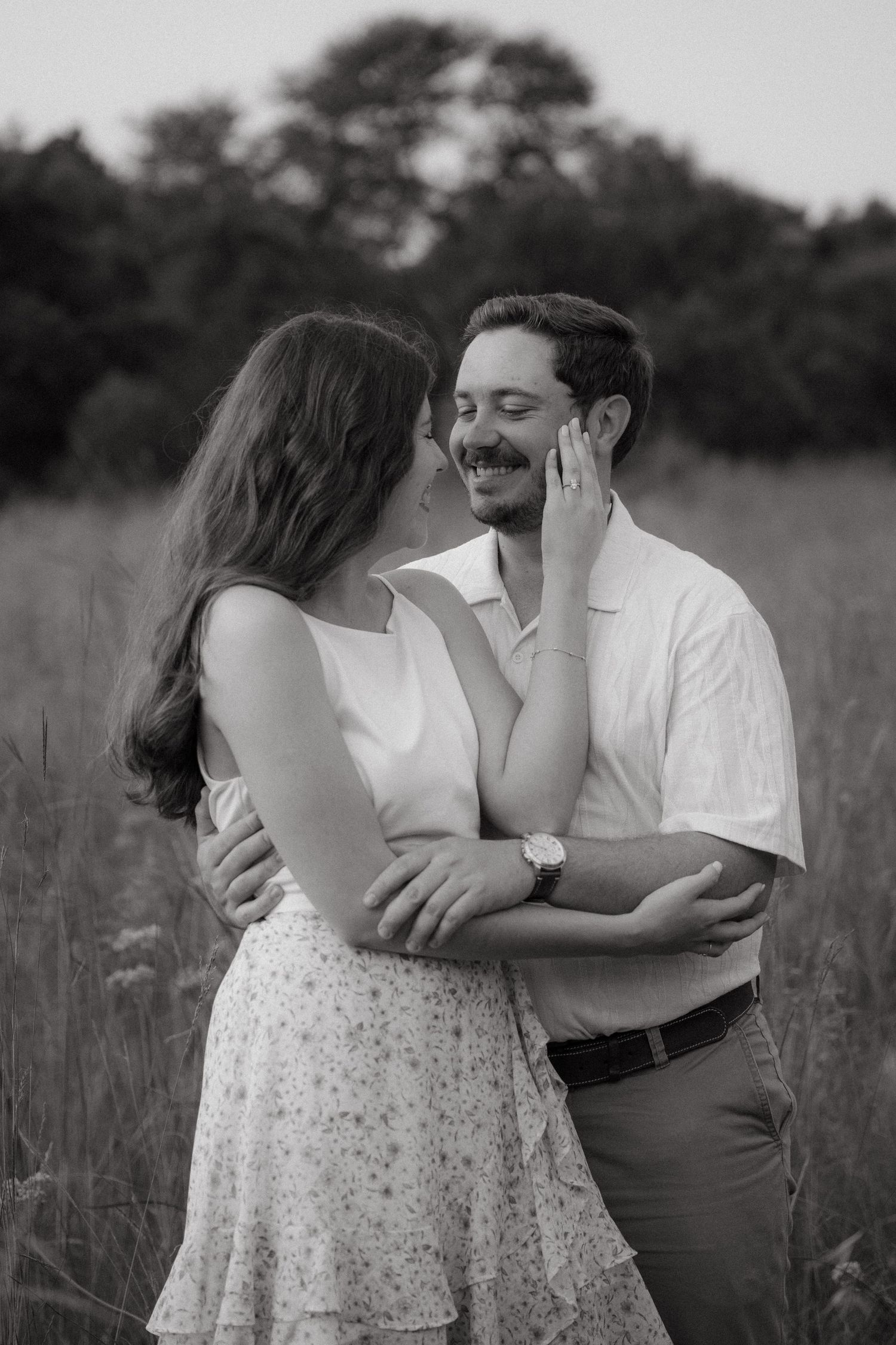 Black and white photo of a smiling couple embracing in a field, woman touching mans face tenderly.
