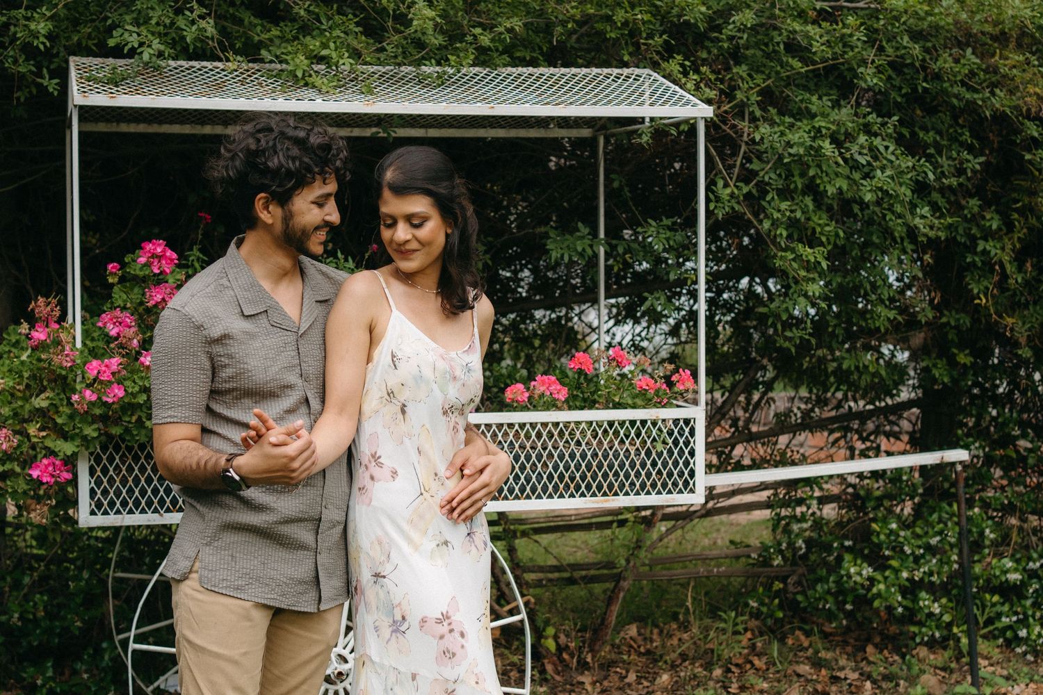 Couple posing together by a garden gazebo with pink flowers blooming in background.
