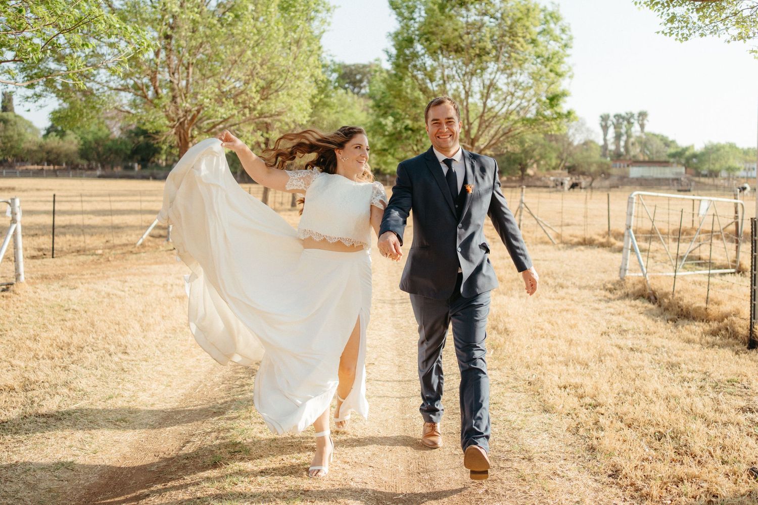 A wedding couple playfully walking through a sun-dried field with the bride's dress flowing in the wind.