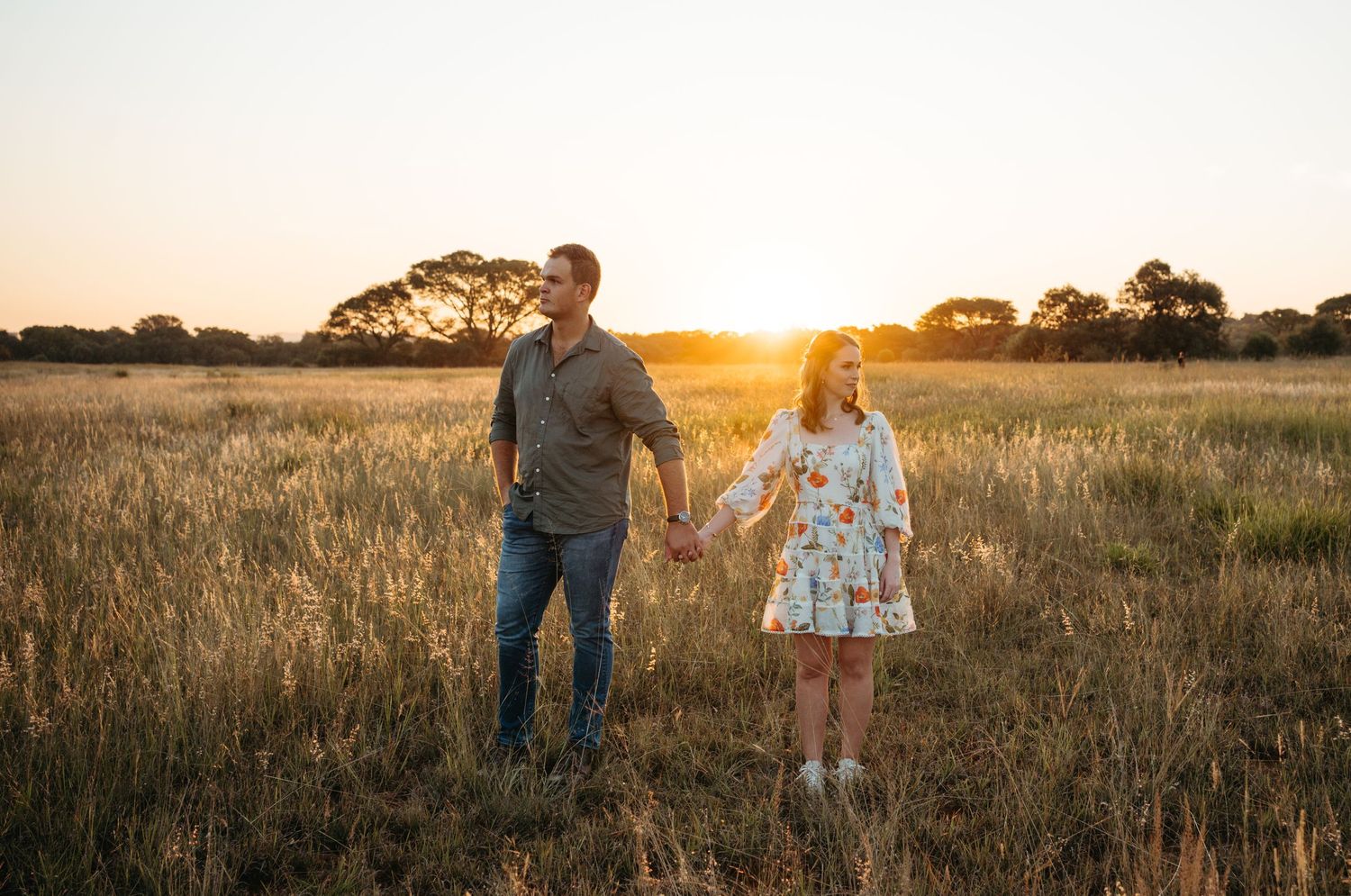 A couple standing together in a golden field at sunset with trees in the background.