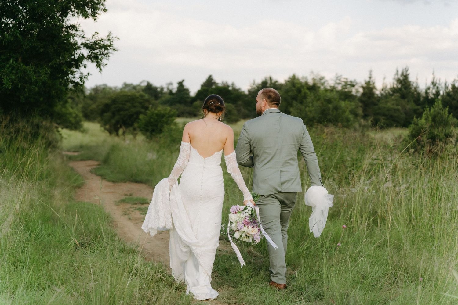 A newlywed couple walks hand in hand through tall grass in a rustic outdoor setting.