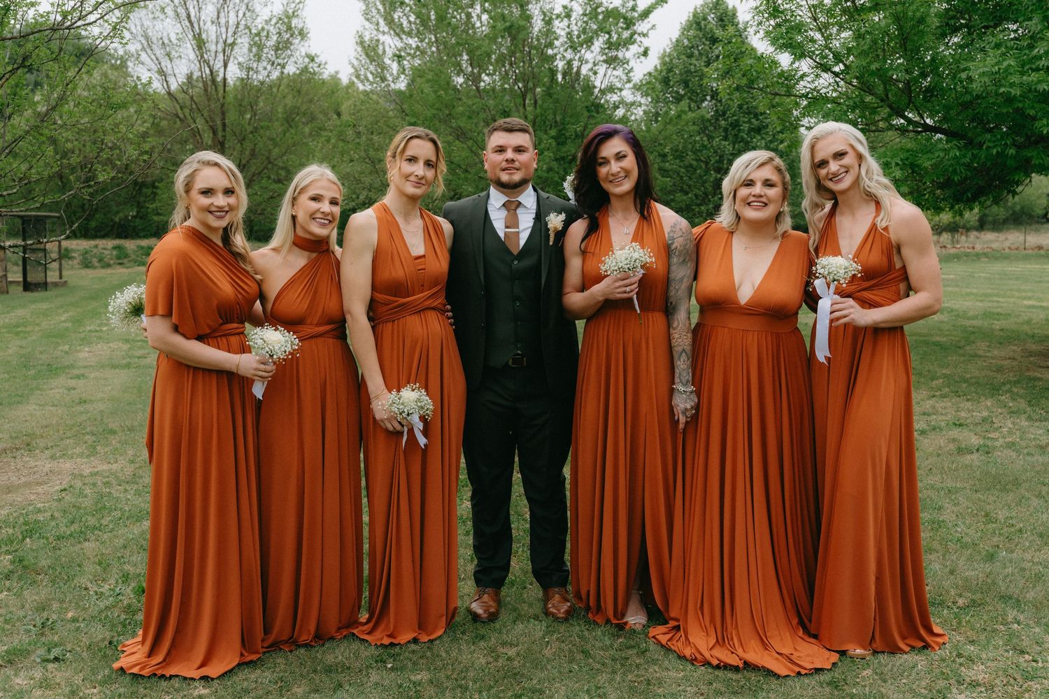 Bridal party in burnt orange dresses poses outdoors for wedding photos.