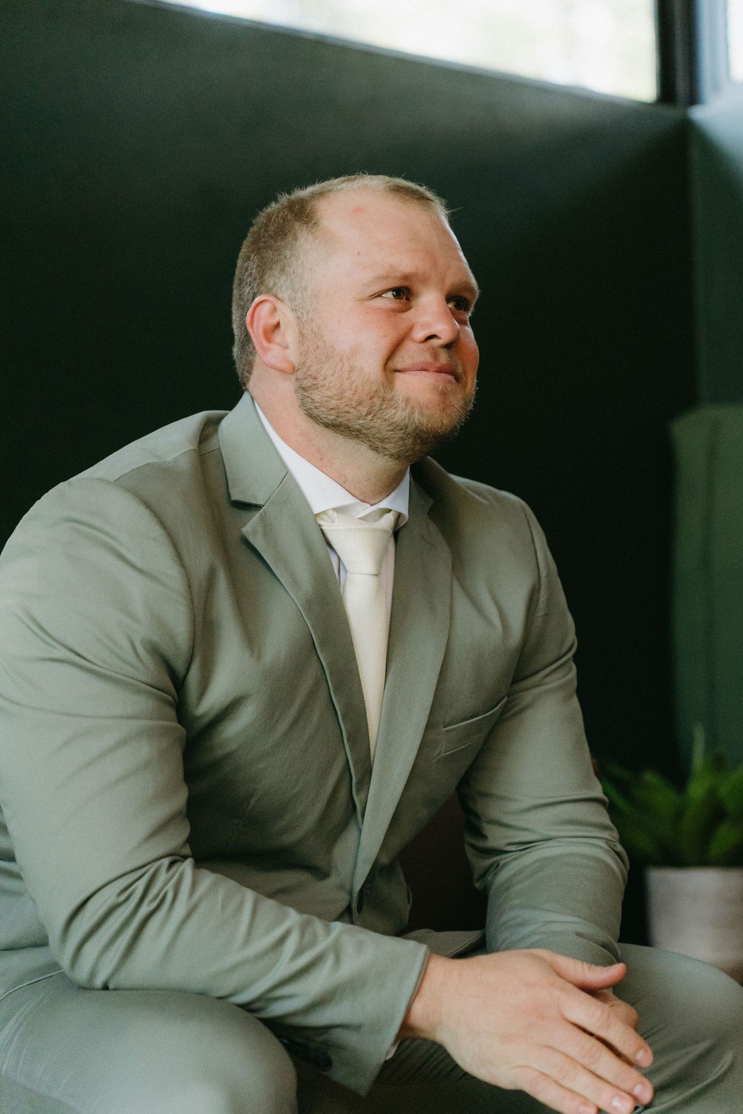A person in a light gray suit and white tie smiles while seated in an indoor setting with natural lighting.