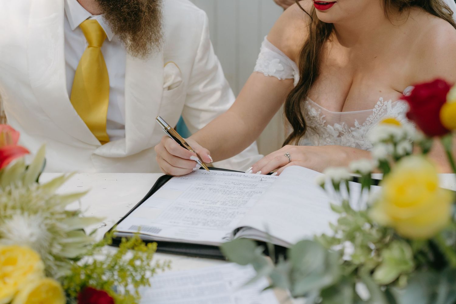 Wedding couple signing marriage certificate with colorful floral arrangements on table.