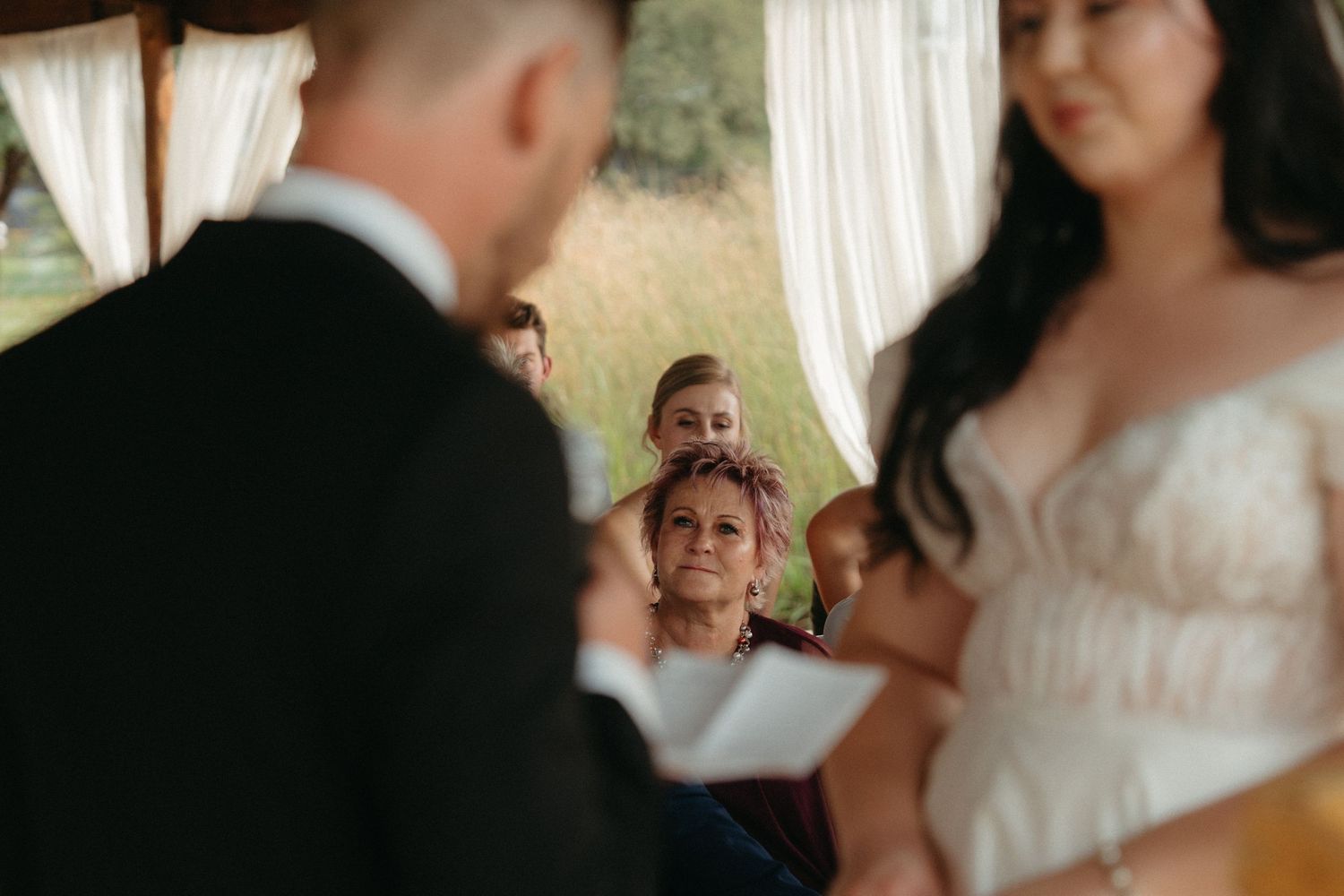 A wedding ceremony moment captured through sheer white curtains in an outdoor setting.