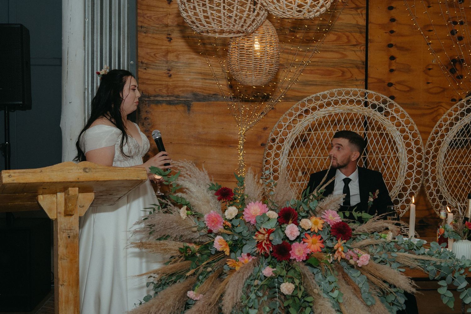 Wedding reception setup with peacock chairs, floral arrangements, and rustic wooden backdrop.