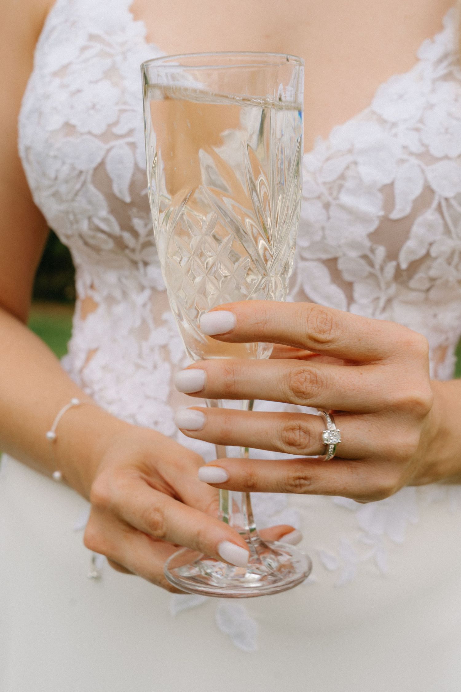 Close-up of hands holding a crystal champagne glass, showcasing an elegant engagement ring and lace wedding dress.