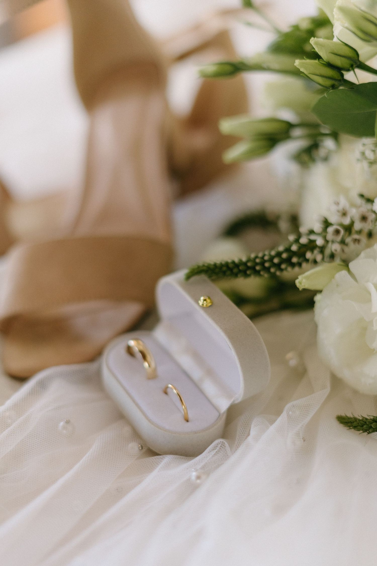 Wedding rings nestled in a white box alongside delicate white flowers and greenery on soft fabric.