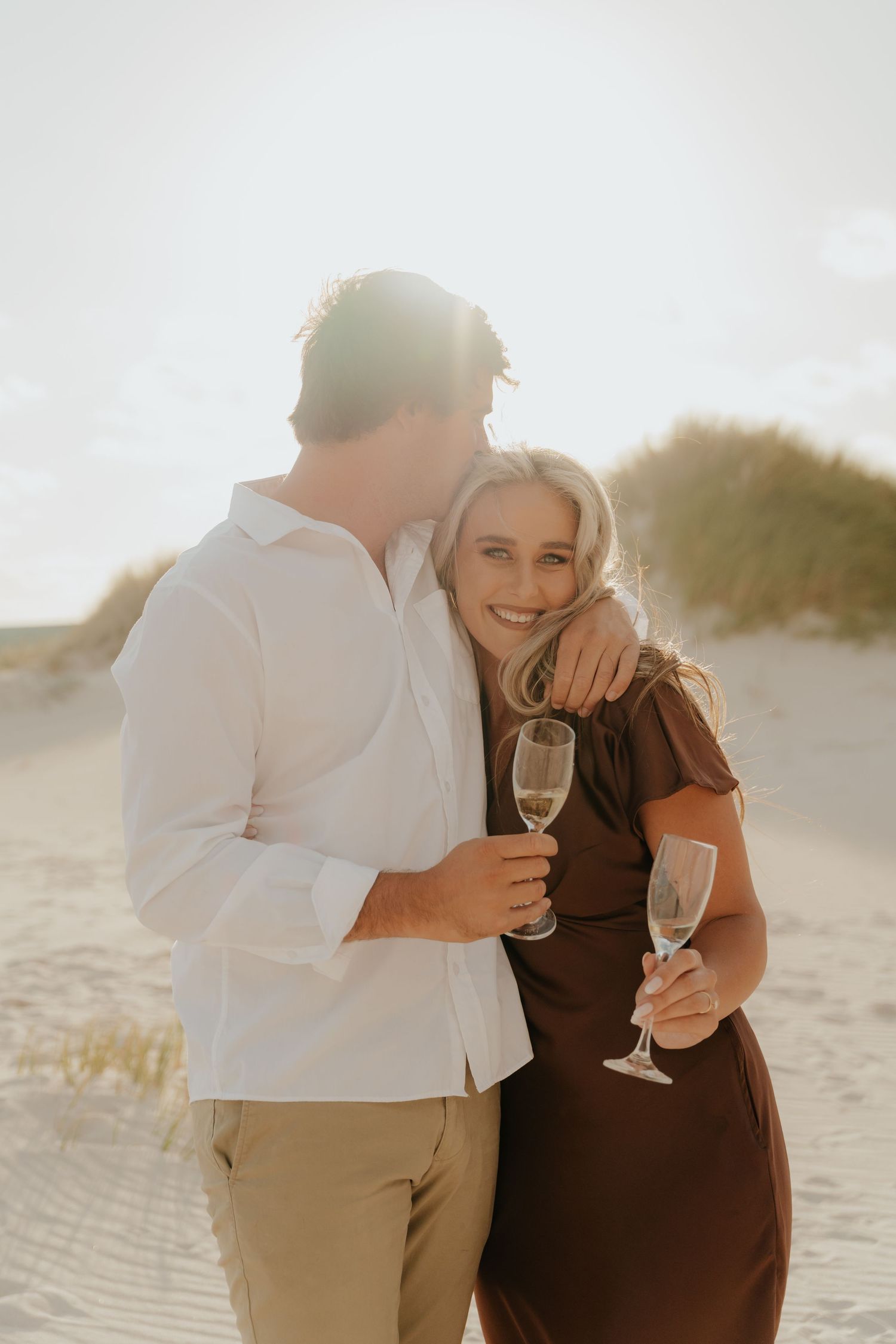 Couple celebrating on sandy beach at sunset, holding champagne glasses and smiling.