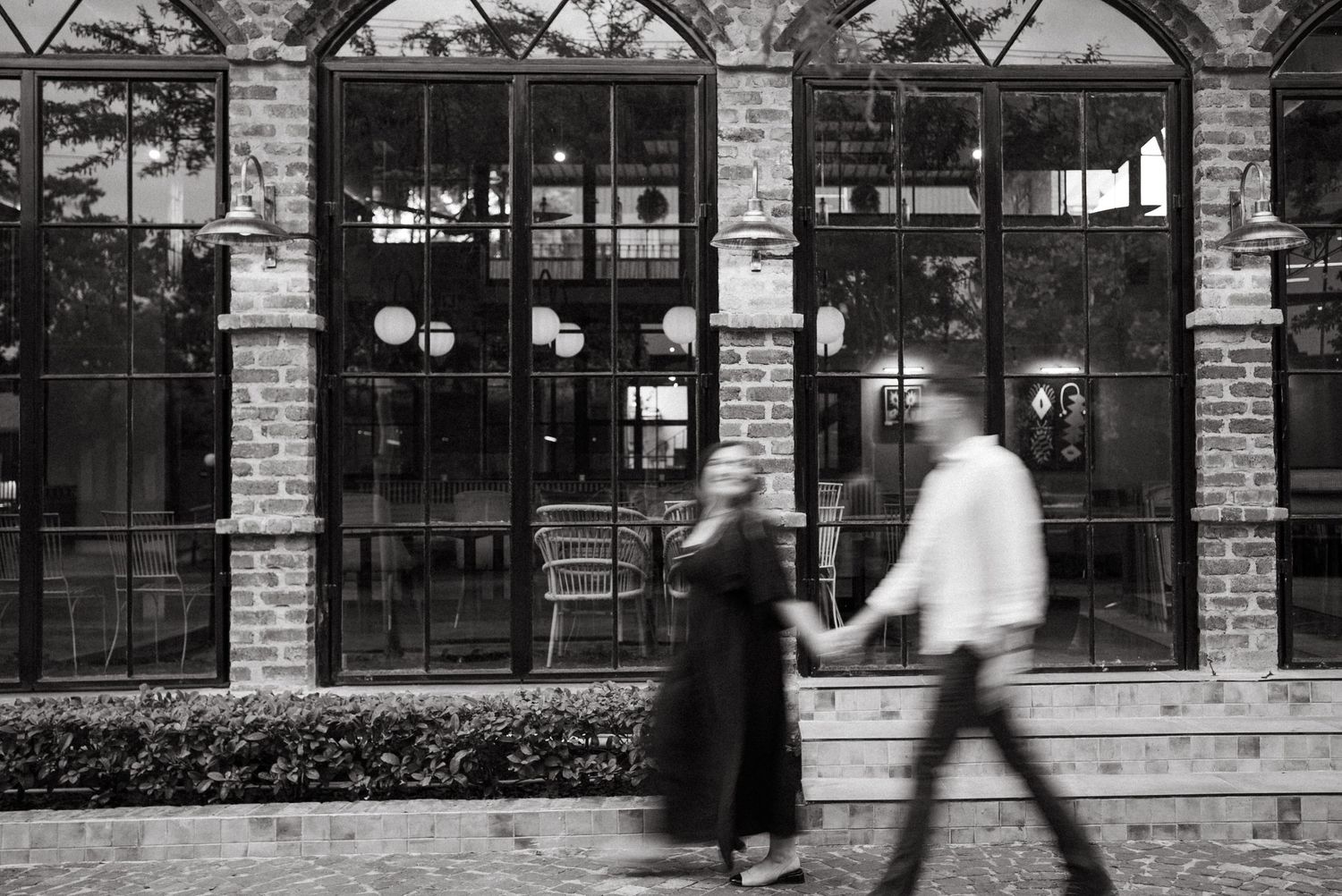 Motion-blurred figures pass by vintage arched windows of a brick building in black and white photography.