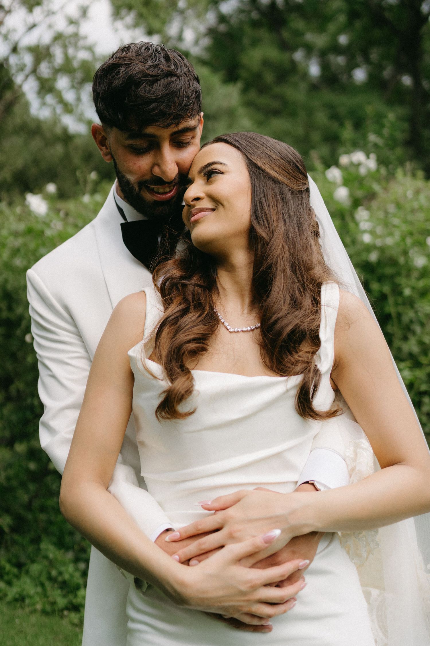 A romantic wedding portrait of a couple embracing in a garden setting with greenery and wildflowers in the background.