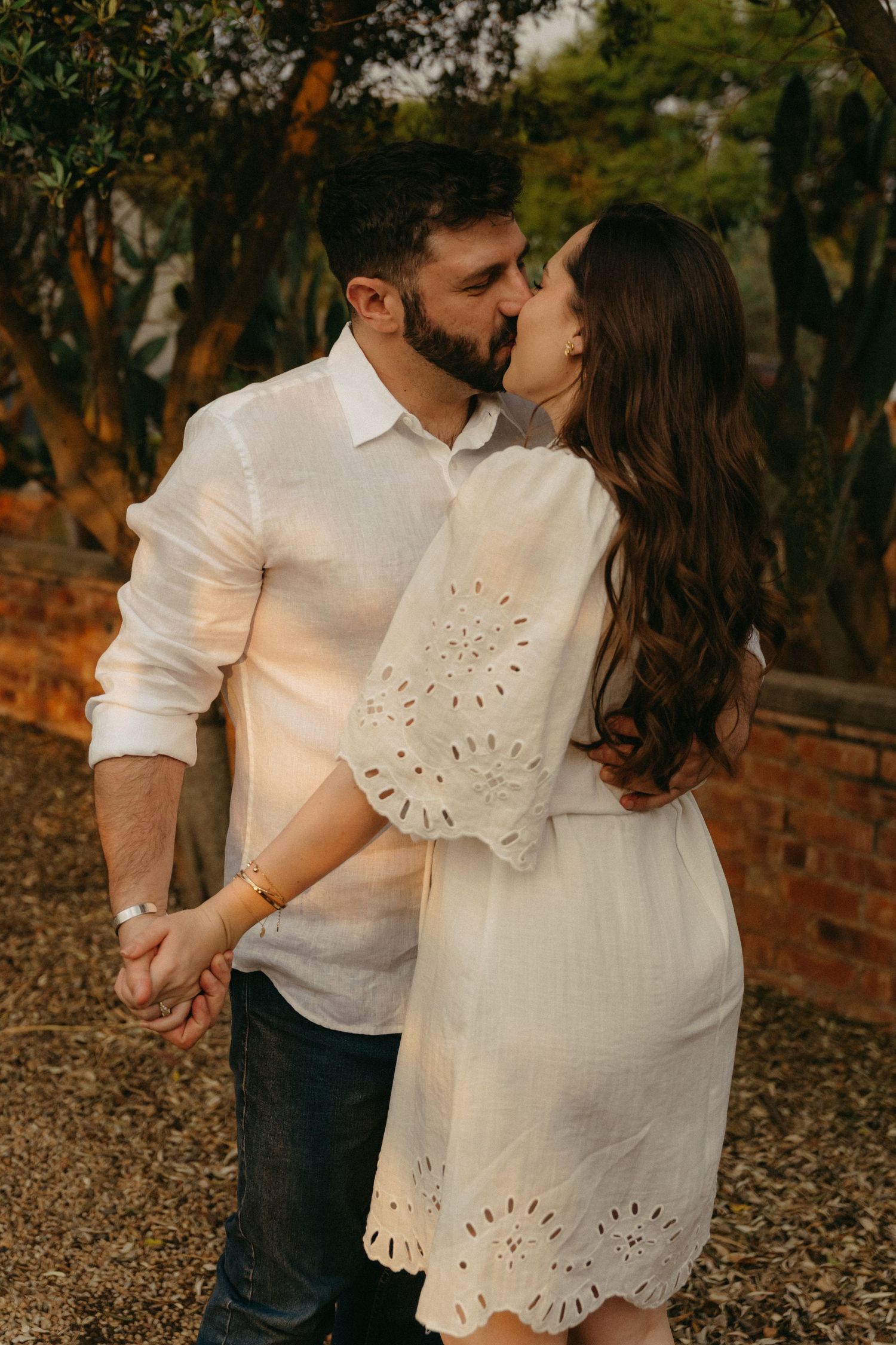 Romantic couple in white clothing sharing tender moment against brick wall in garden setting.
