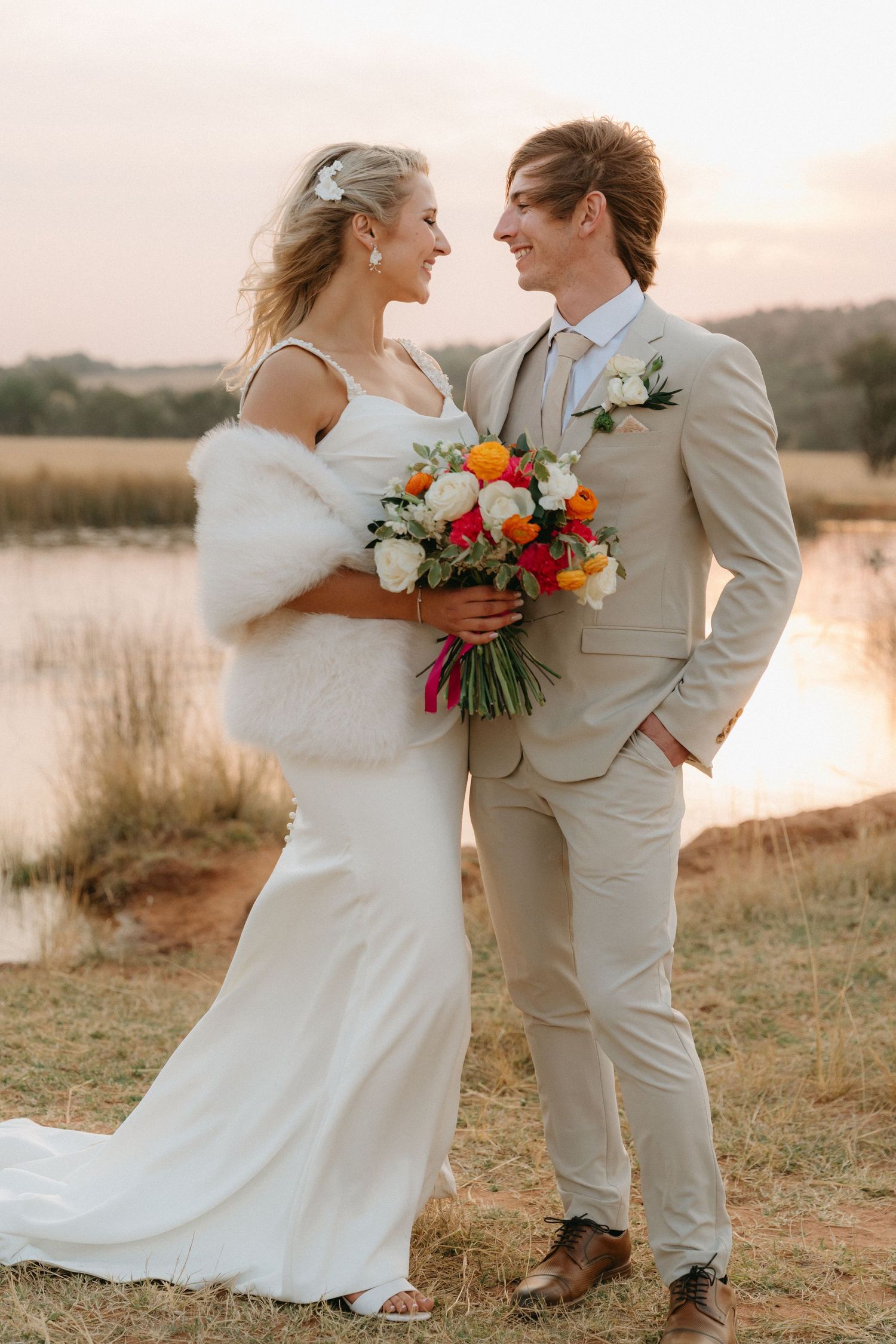 A romantic wedding couple embraces in a field at sunset with colorful bouquet details.