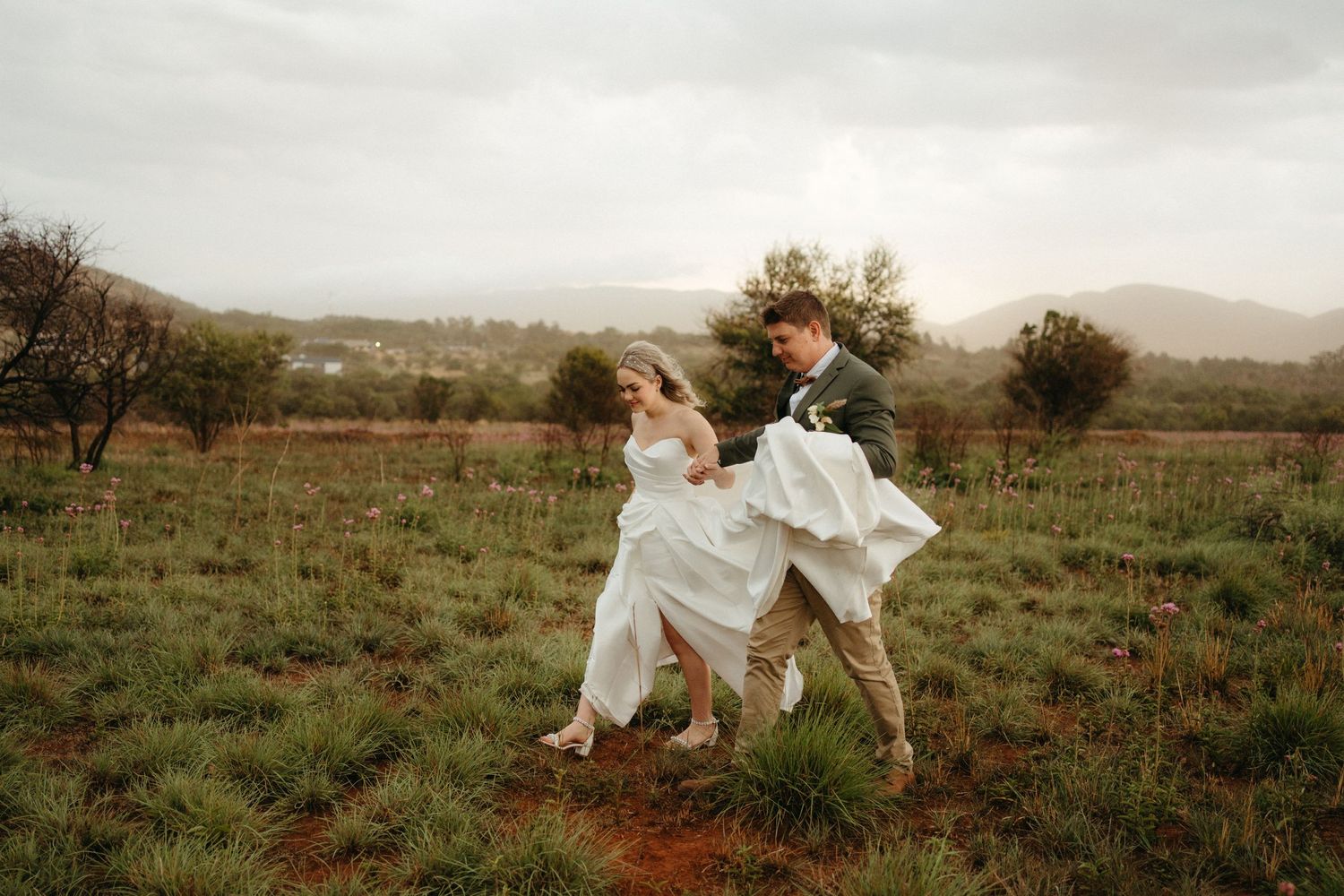 A couple dances joyfully in a rustic field with flowing white dress against a scenic countryside backdrop.