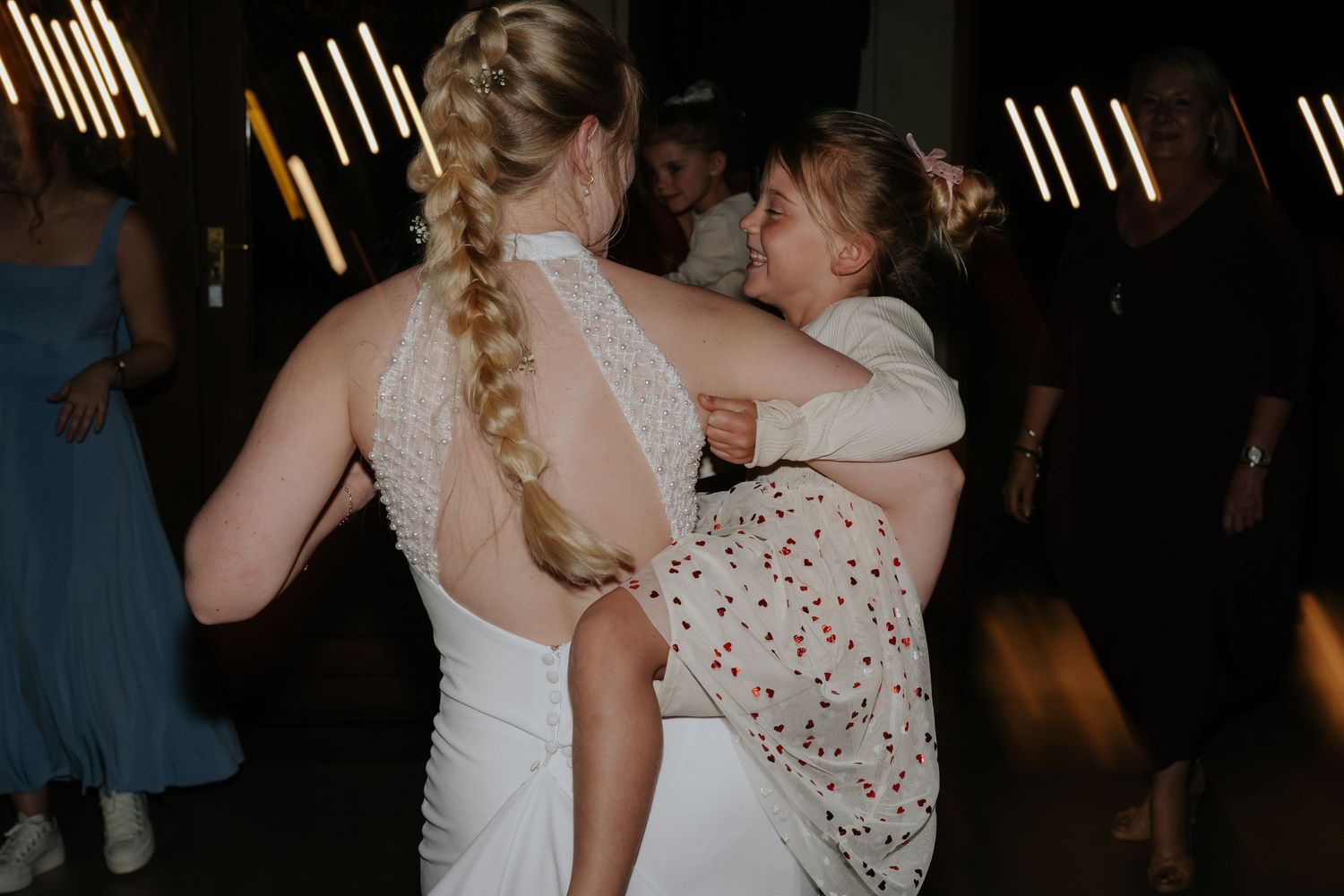 Wedding party dancing and celebrating on a dimly lit dance floor with festive lighting overhead.