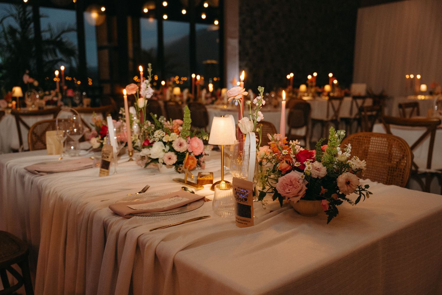 Elegant wedding reception table with floral centerpieces, candles, and soft pink linens in a warmly lit venue.