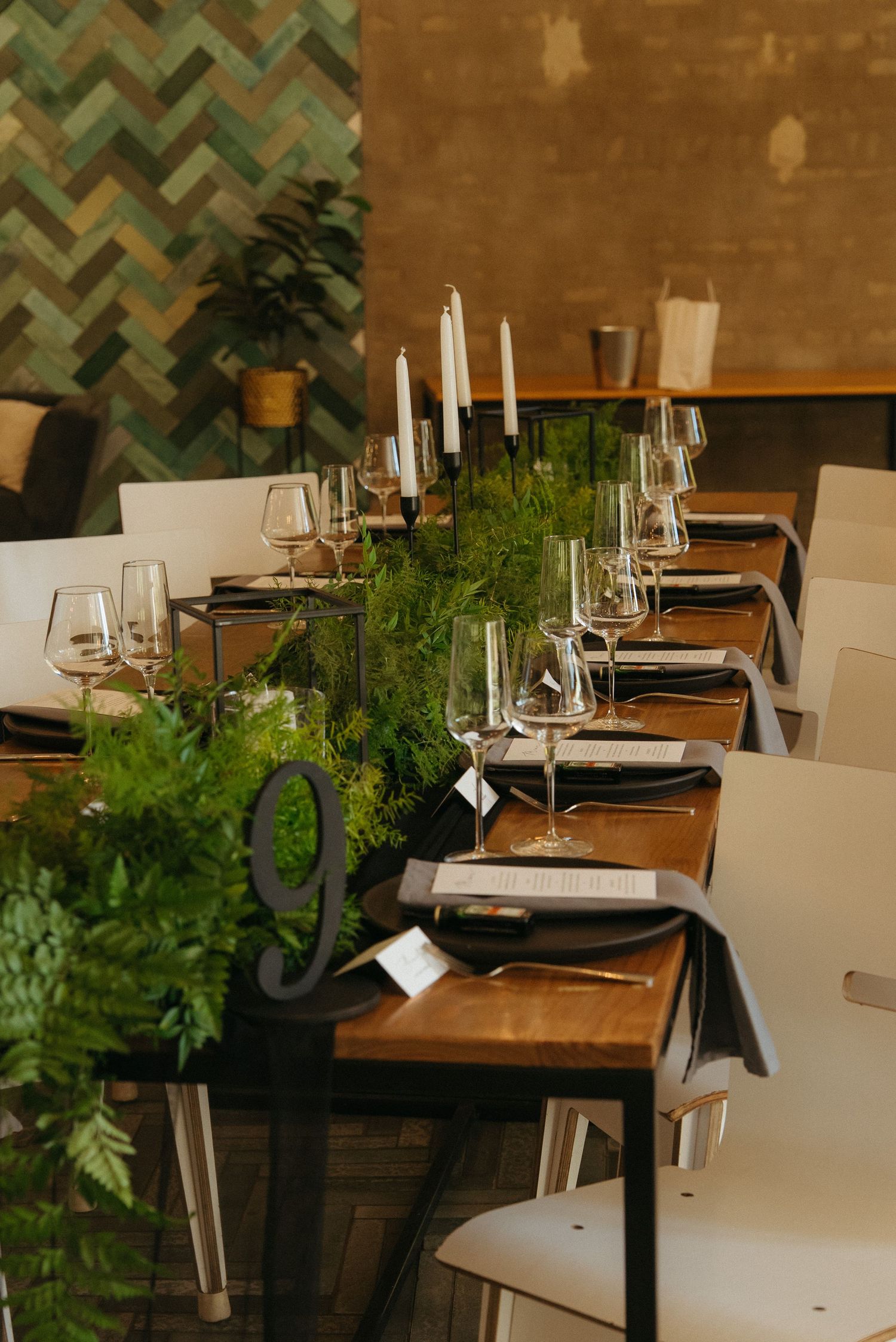 Elegant table set with greenery runner, white candles, wine glasses, and black napkins on wooden tables with white chairs.