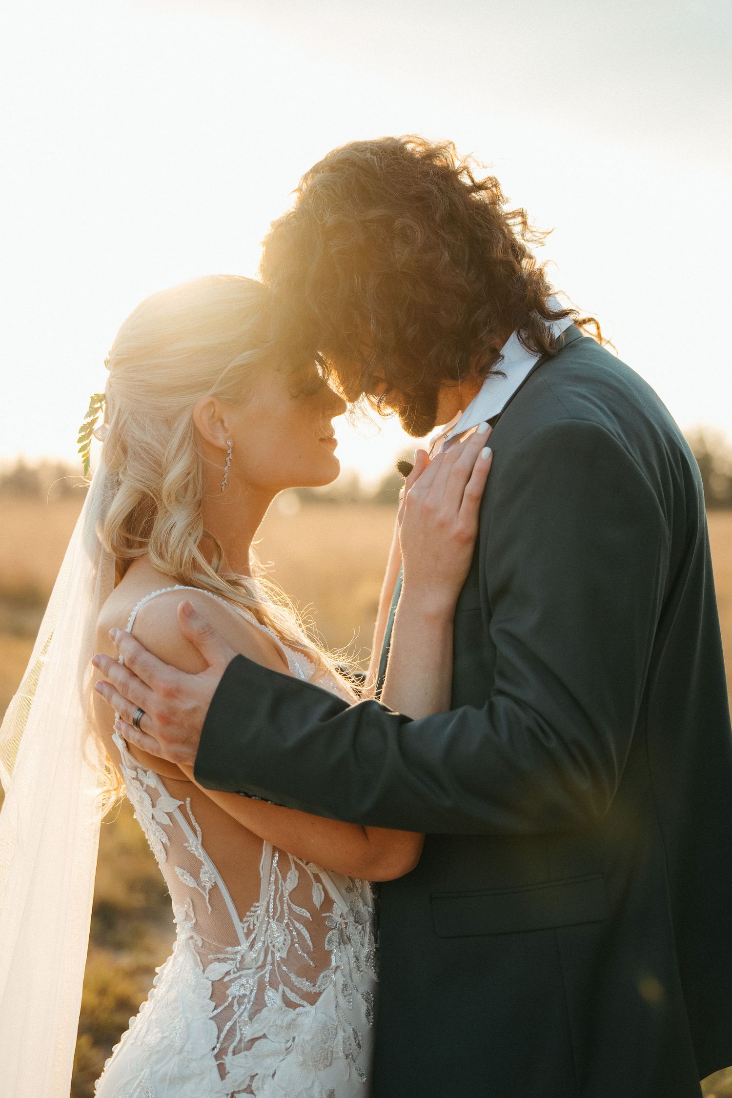 A bride and groom share a romantic golden hour moment in a sunlit field, with the bride in lace and groom in a dark suit.