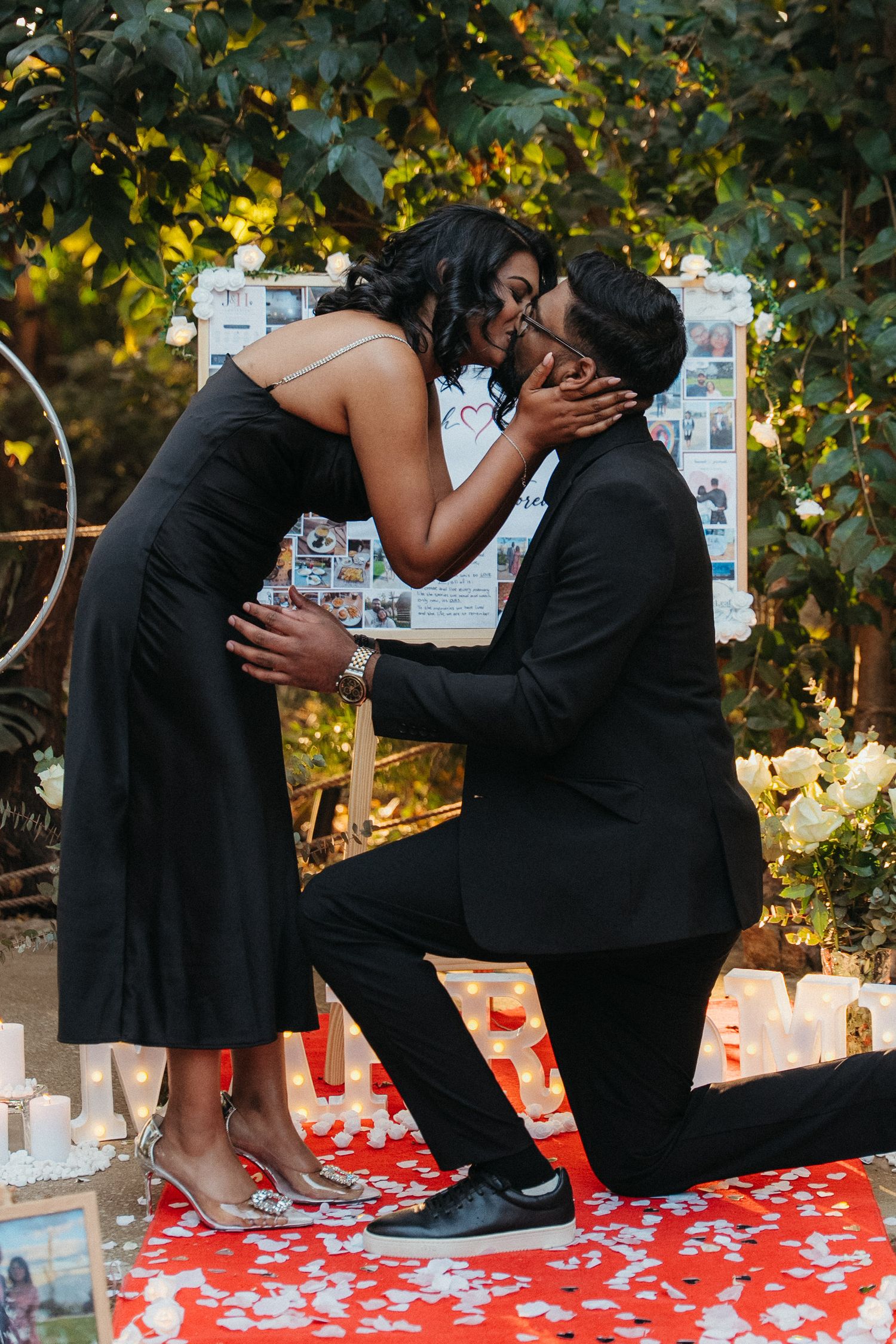 Couple sharing a kiss during a romantic outdoor proposal with candles, rose petals, and a photo board backdrop.