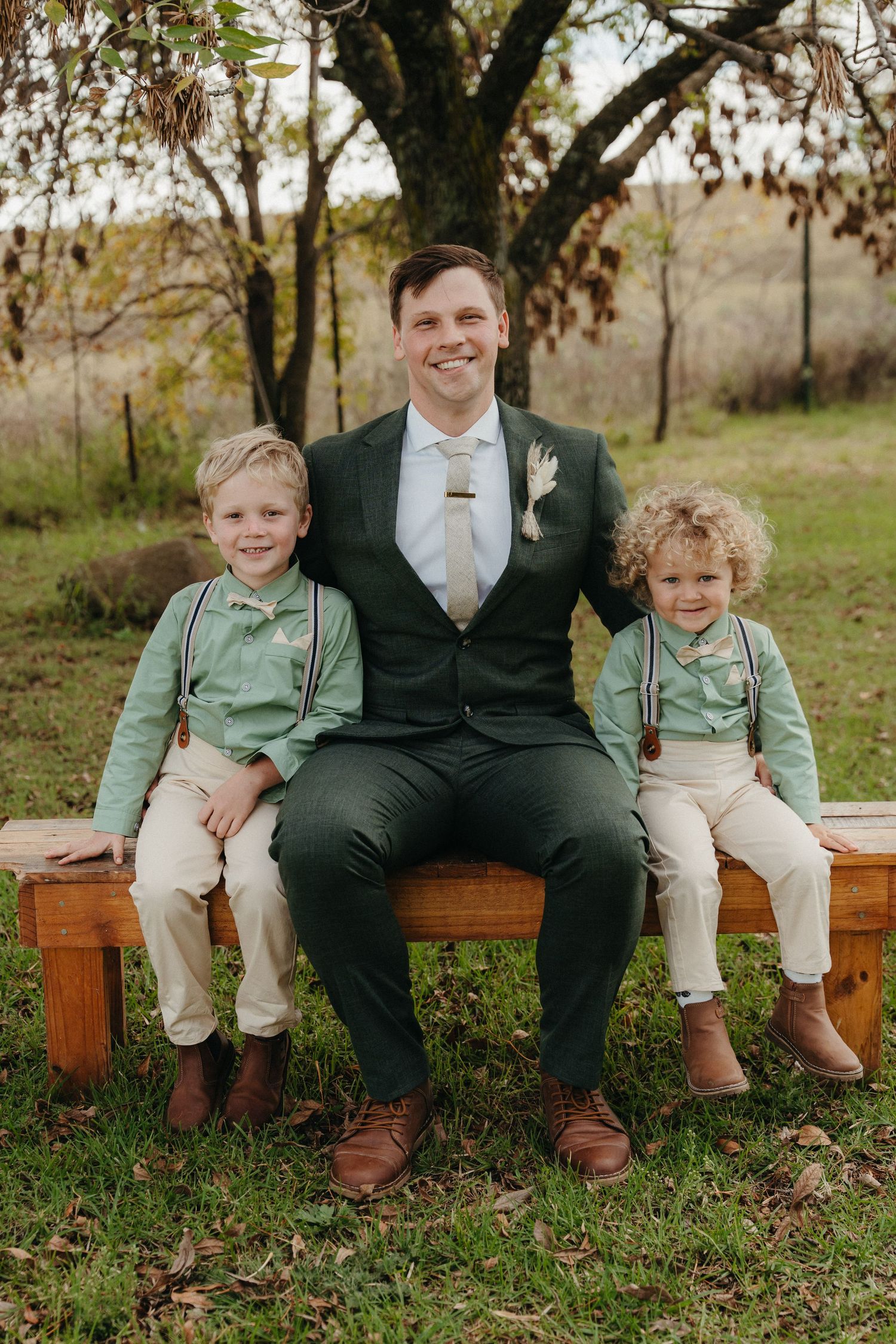 A groom in a dark green suit sits on a wooden bench with two young boys in matching sage suspenders at an outdoor wedding.