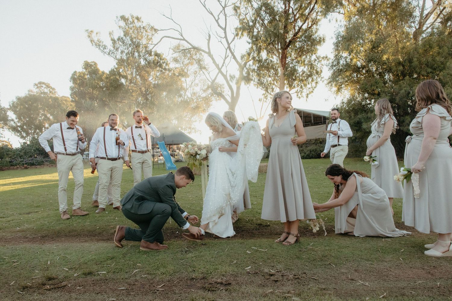 Wedding party gathered outdoors on grass, bride and groom kneeling with guests in neutral attire during golden hour ceremony.
